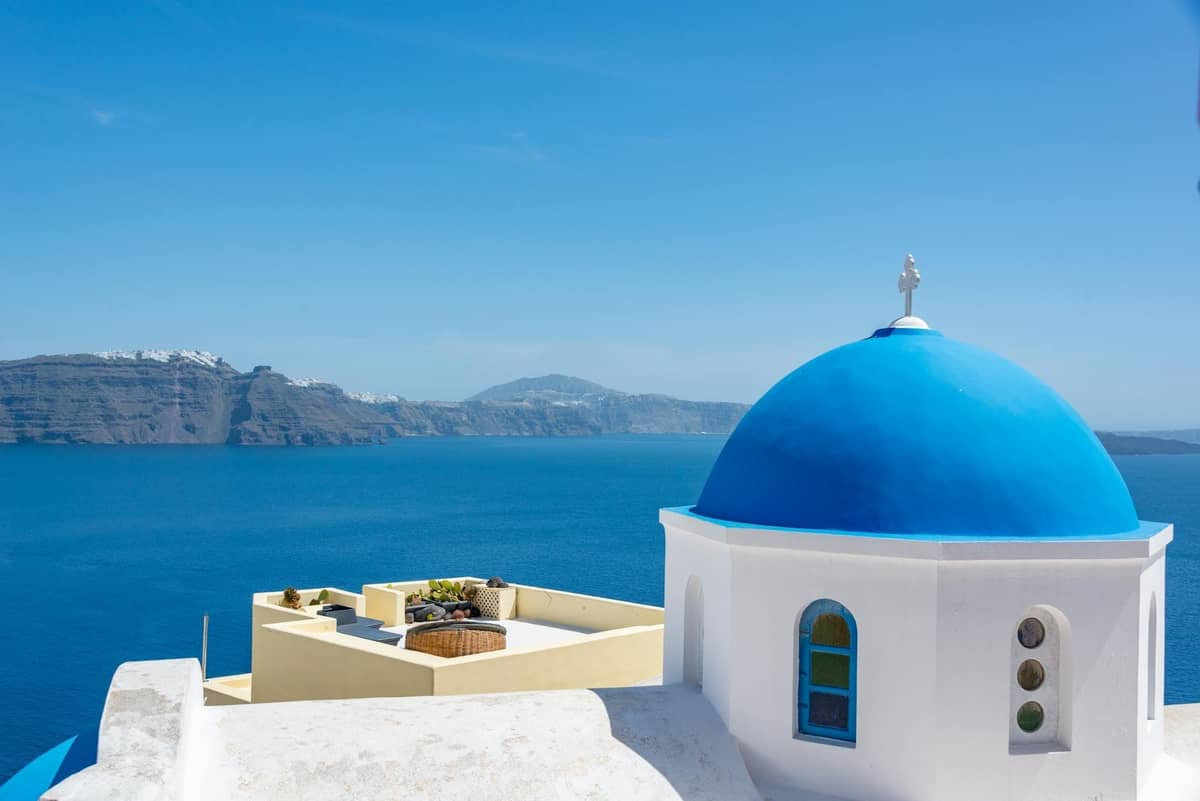 Iglesia de cúpula azul en Santorini con la caldera y la isla de Thirasia visible al otro lado del mar en un día despejado
