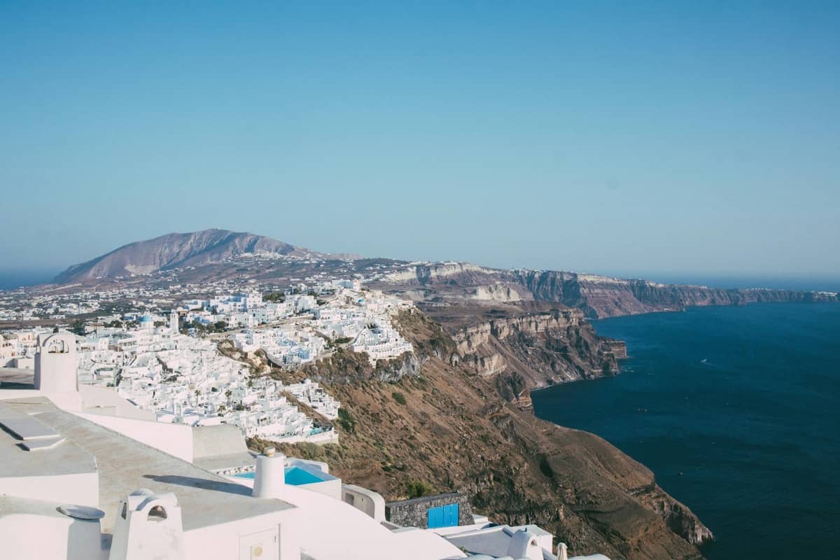 Vista amplia del borde de la caldera de Santorini con edificios encalados en los acantilados, el mar azul abajo y la famosa ruta a pie de Fira a Oia visible.