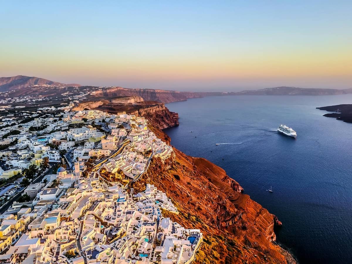 Widok z lotu ptaka na klify kaldery Santorini otoczone luksusowymi hotelami i willami pobielanymi wapnem z widokiem na głębokoniebieskie morze i statek wycieczkowy o zachodzie słońca.