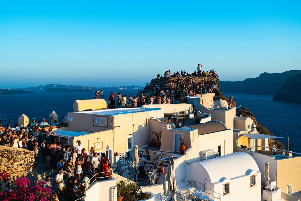 Duży tłum ludzi zebranych na dachach w Oia, Santorini, czekających na zachód słońca z widokiem na kalderę w tle.