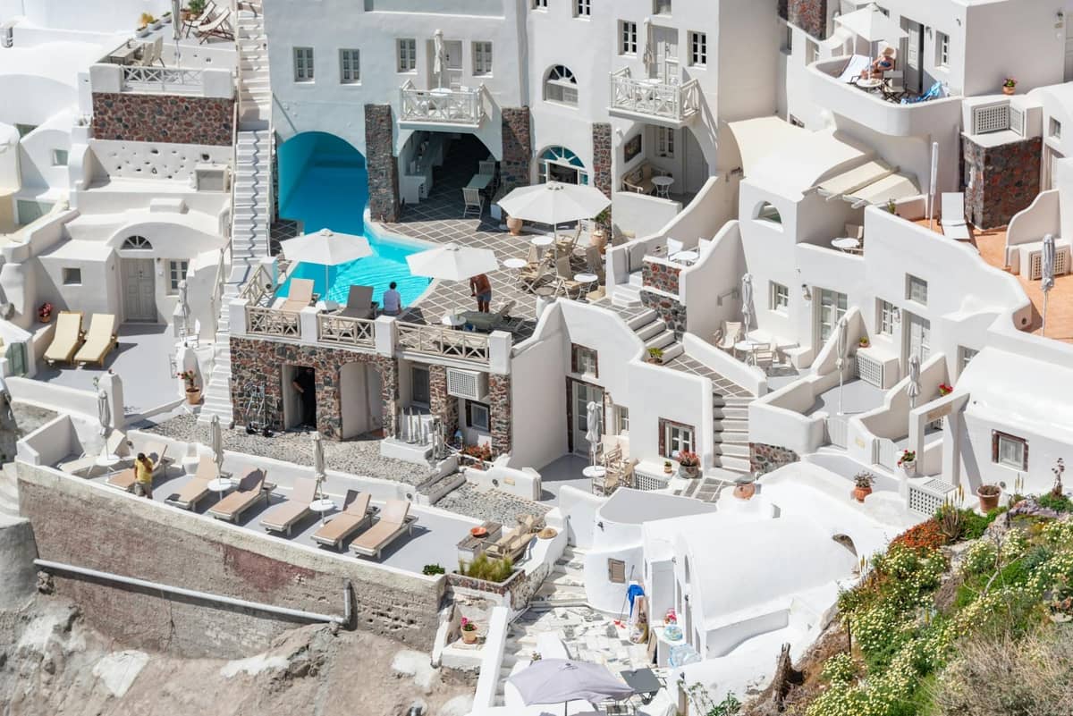 Un conjunto de alojamientos encalados de estilo clásico en Santorini con balcones, tumbonas y una piscina, construidos en el acantilado bajo la brillante luz del sol.