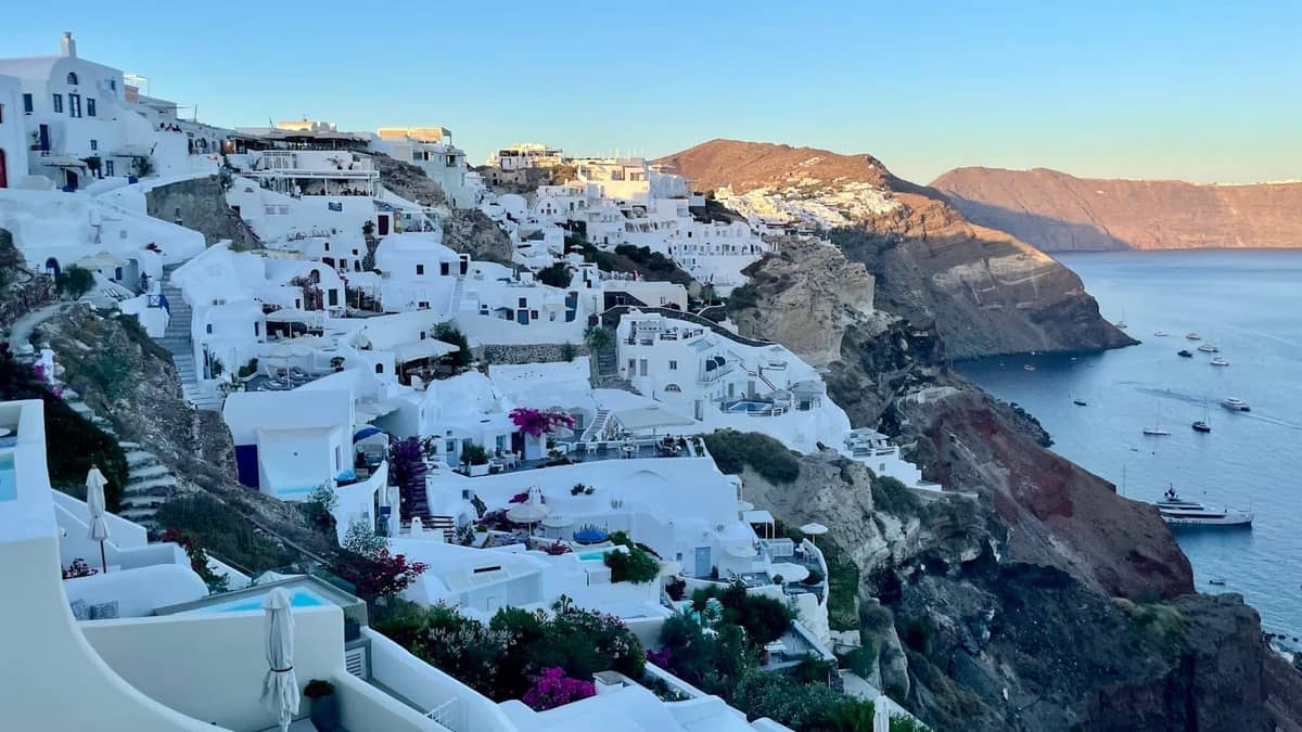 Vista de los edificios blancos de Santorini en el acantilado con vistas al mar azul, cielo despejado, luz del amanecer y poca gente.