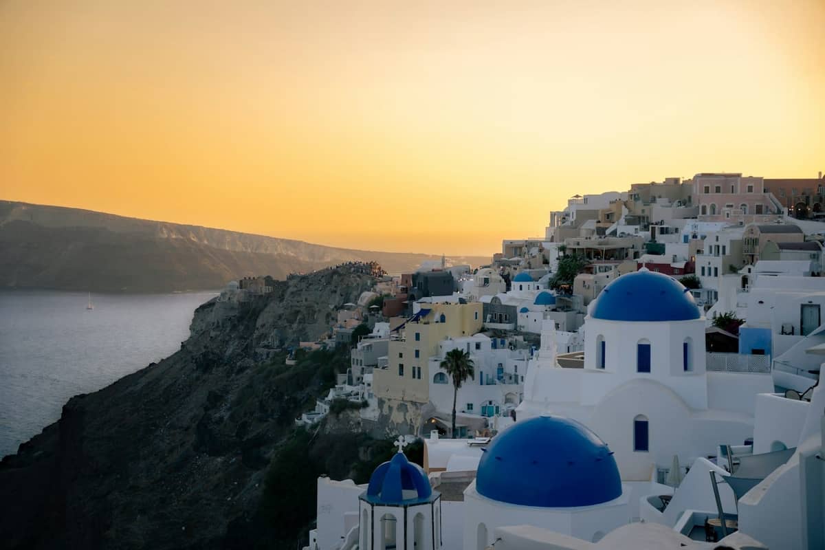 Borde de la caldera de Santorini con las icónicas iglesias de cúpula azul, edificios encalados en los acantilados y un cielo dorado al atardecer sobre el Mar Egeo.