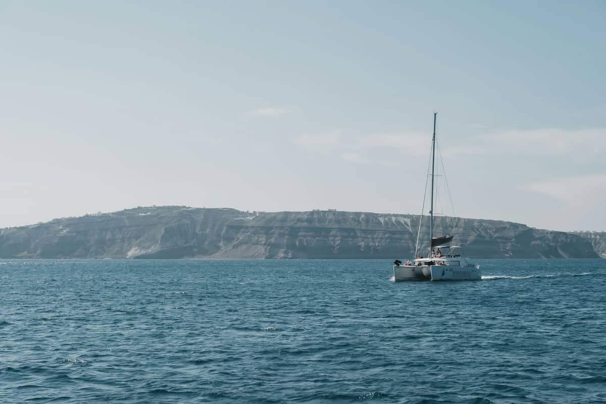 Katamaran segelt nahe den markanten Klippen von Santorini unter einem klaren Himmel, mit der Küstenlinie der Insel in der Ferne.