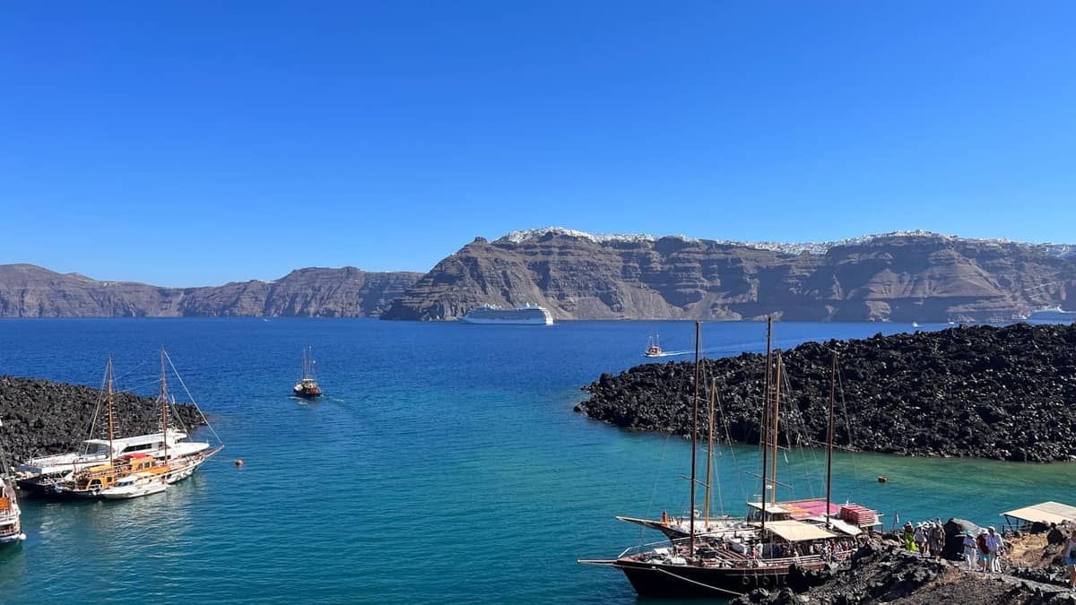 Santorini-Kaldera-Marina mit traditionellen Segelbooten, an Bord gehenden Personen und Vulkanklippen im Hintergrund unter einem klaren blauen Himmel.