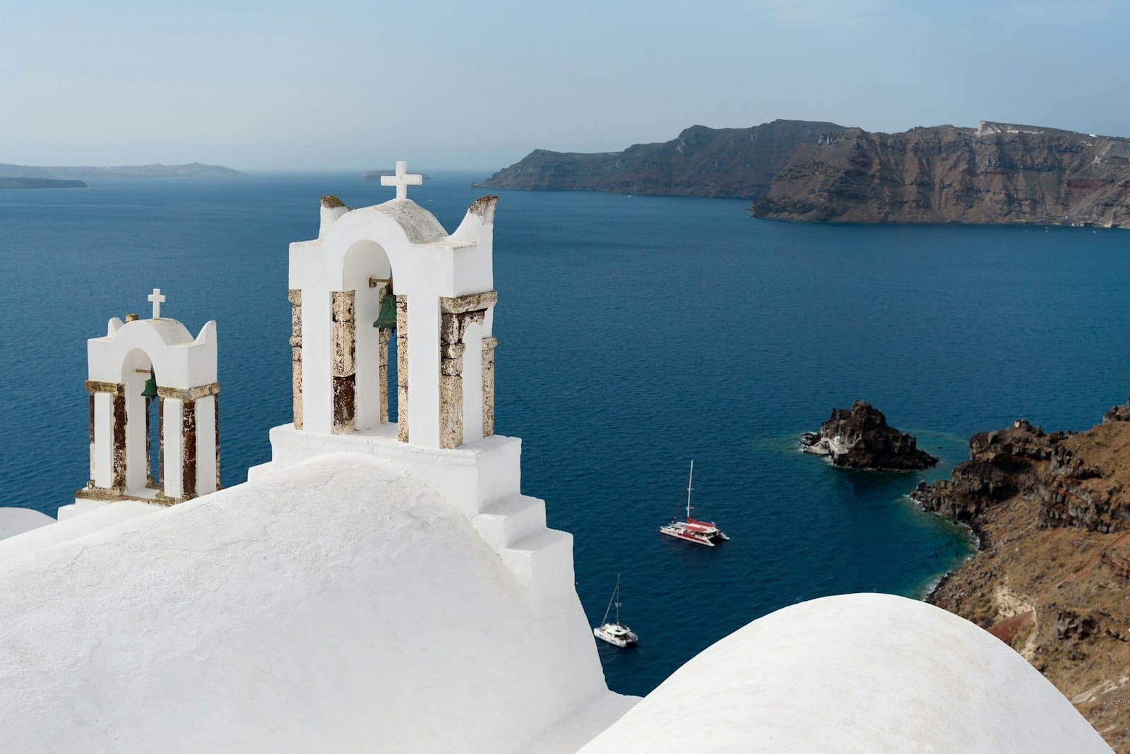 Sinos de igreja brancos no terraço com vista para o Mar Egeu azul, catamarãs navegando abaixo e as falésias da caldeira de Santorini ao fundo.