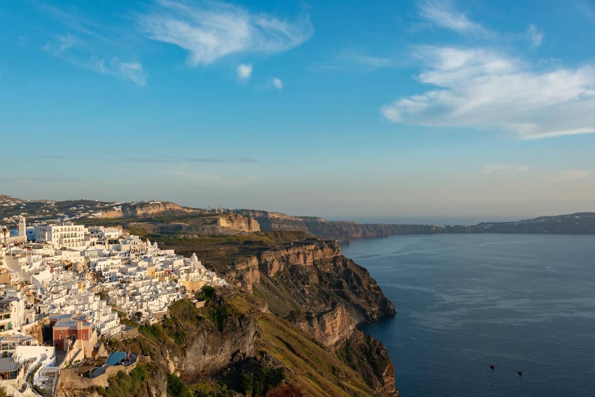 Vista del borde de la caldera de Santorini con edificios blancos encaramados en dramáticos acantilados con vistas al Mar Egeo bajo un cielo azul.