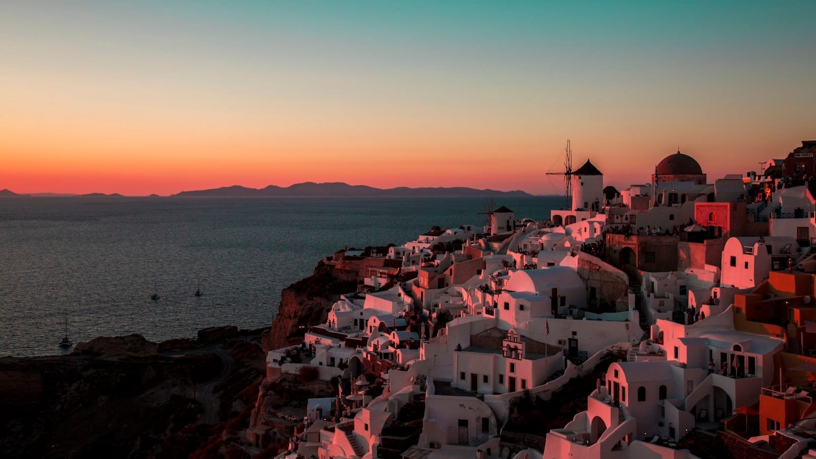 Os icônicos prédios brancos com cúpulas azuis de Santorini com vista para o mar ao pôr do sol, com cores vibrantes de laranja e rosa no céu e a silhueta clássica de um moinho de vento.