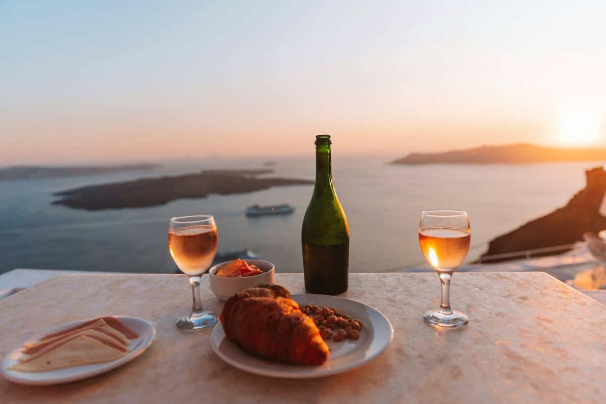 Una mesa con vino, copas y comida con vistas a la caldera de Santorini al atardecer, con luz dorada sobre el mar y los acantilados.