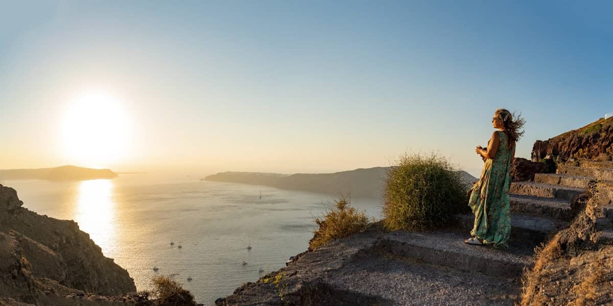 Mujer de pie en un camino con vistas a la caldera de Santorini al atardecer, con amplias vistas y luz dorada de la tarde