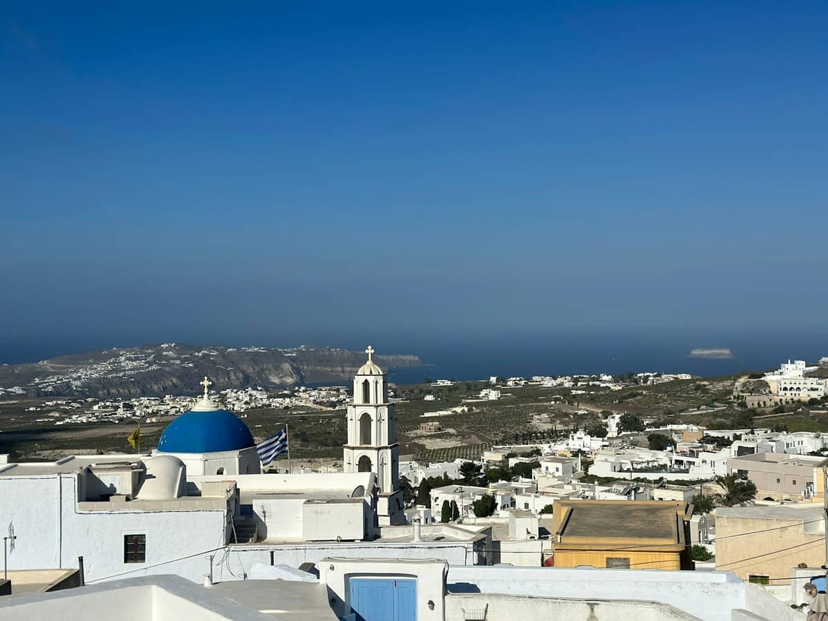 Vista panoramica da un villaggio dell'entroterra di Santorini con edifici bianchi, una chiesa con cupola blu e ampie vedute sull'isola e sul mare.