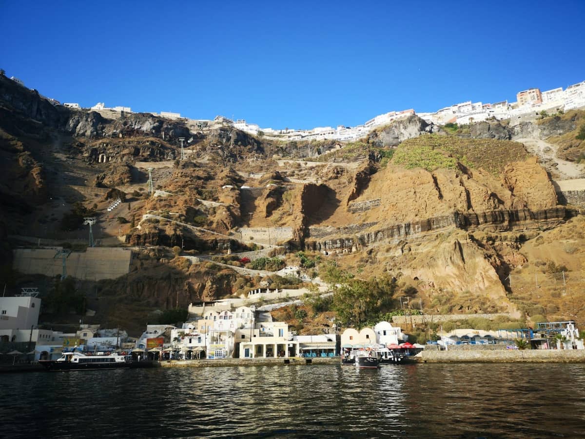 Vista dal livello dell'acqua delle scogliere della caldera di Santorini con edifici bianchi arroccati in alto, porto dei traghetti e piccole barche alla base.