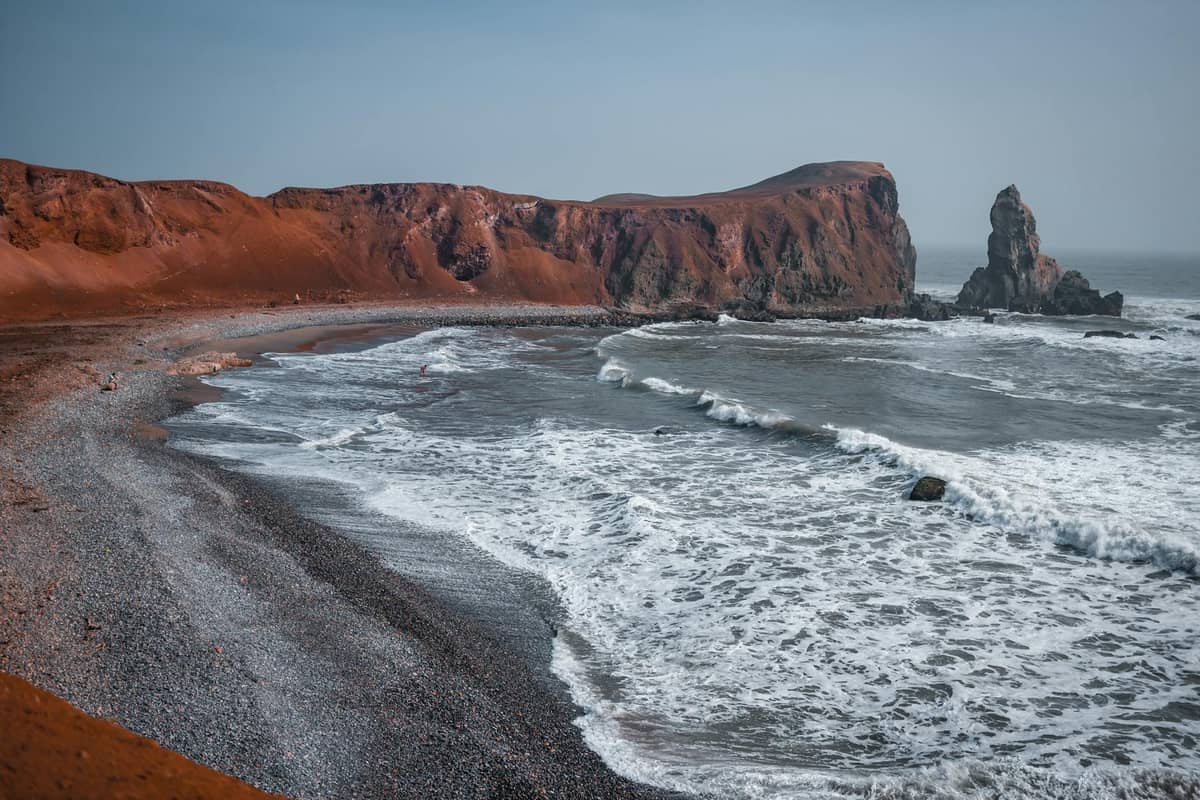 Vista panoramica di una drammatica costa con scogliere rosse, onde che si infrangono su una spiaggia sassosa, un promontorio e uno stack marino a Santorini.