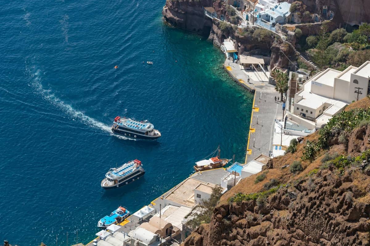 Luftaufnahme des Alten Hafens von Santorini mit angedockten Ausflugsbooten, weißen Gebäuden am Hang, steilen Klippen und türkisfarbenem Meer darunter.