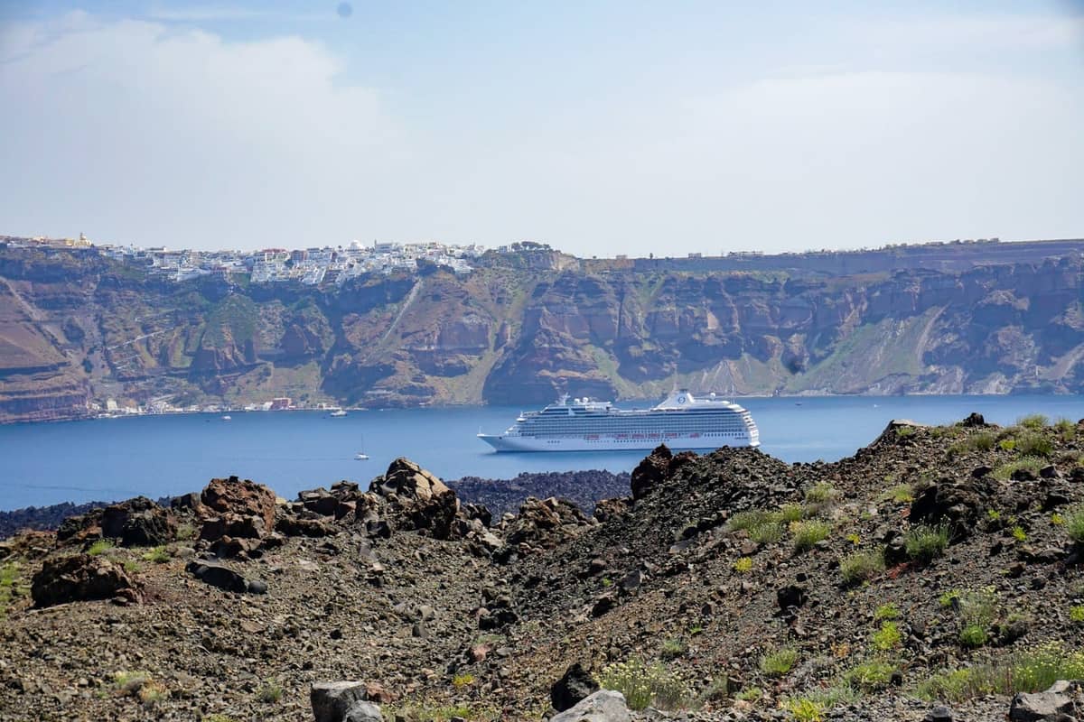 Felsiges Vulkangelände von Nea Kameni mit Kaldera-Klippen und einem Kreuzfahrtschiff im Hintergrund über der Bucht von Santorini