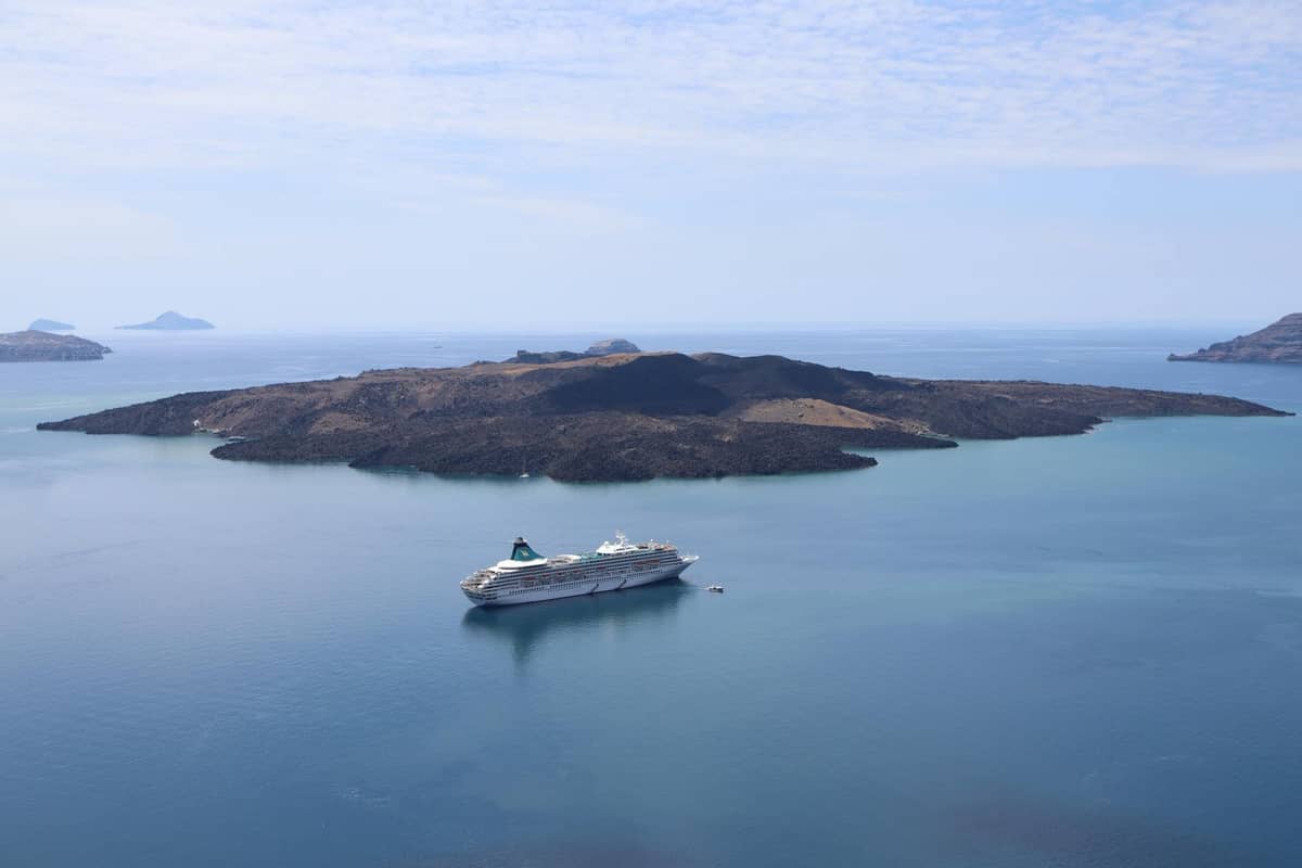Eine Vulkaninsel mit dunklem Felsterrain, umgeben von blauem Meer, mit einem in der Nähe ankernden Kreuzfahrtschiff, das die Caldera von Santorin darstellt.