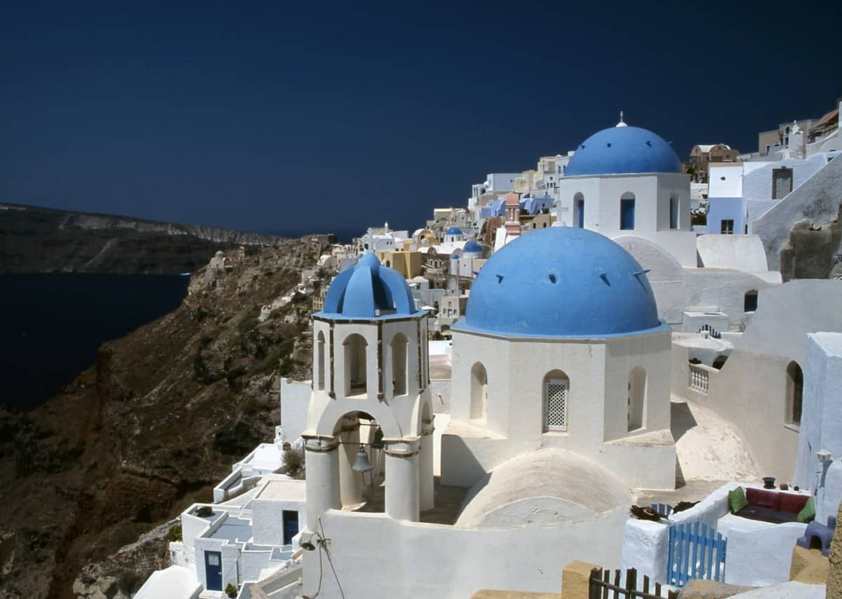 Vista classica della caldera di Santorini con case imbiancate a calce, chiese a cupola blu e scogliere che si affacciano sul mare sotto un cielo sereno.