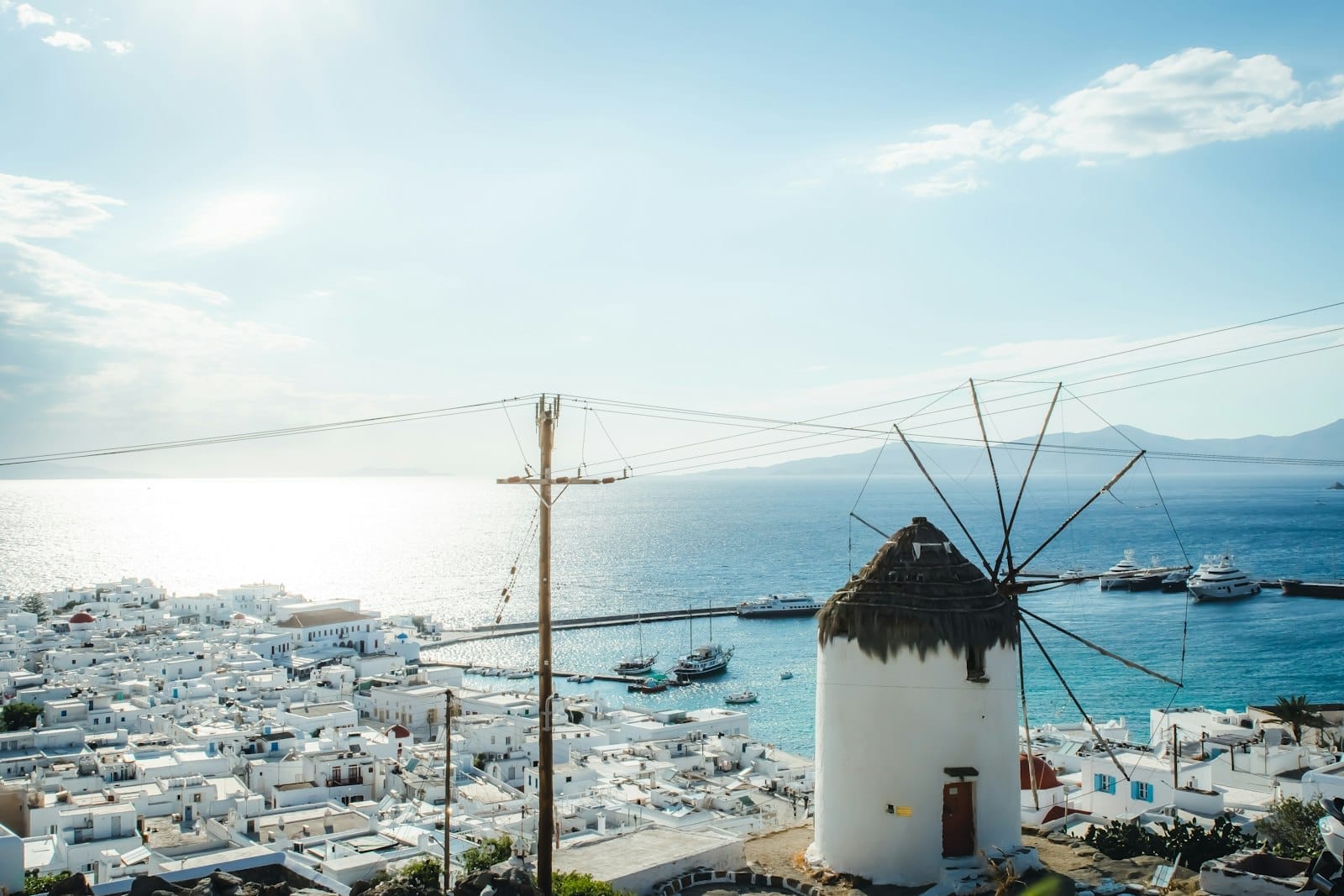 Vista do moinho de vento de Mykonos com o mar azul ao fundo, casinhas brancas e iates ancorados no porto em um dia de sol.