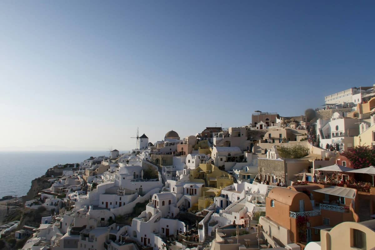 Panoramablick auf Santorinis ikonische weiß getünchte Häuser und blaue Kuppeln an einem sonnigen Tag, mit klarem Himmel und dem im Hintergrund sichtbaren Meer.