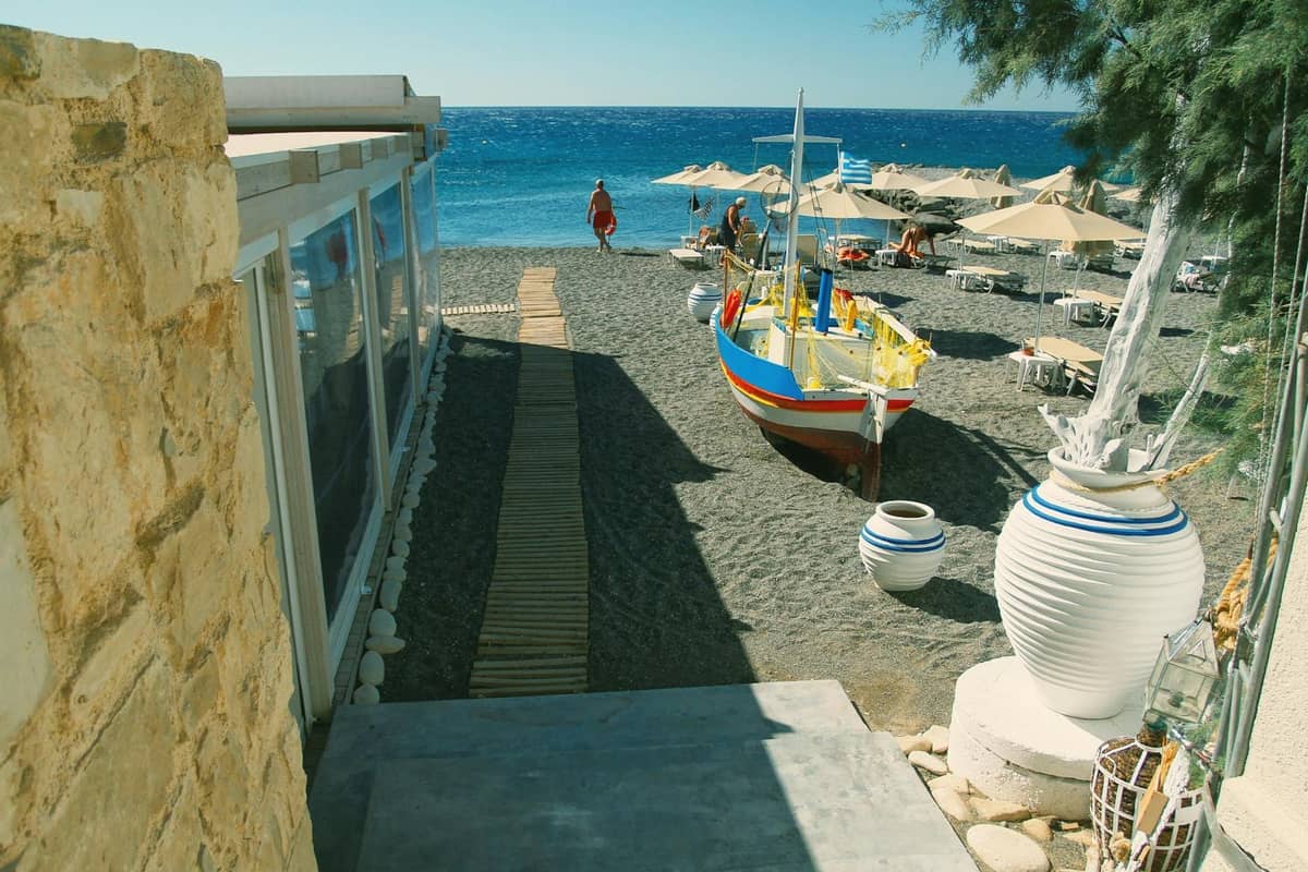 Strandszene auf Santorini mit leuchtend blauem Meer, traditionellem Boot auf dunklem Sand und Liegestühlen unter Sonnenschirmen, die einen mediterranen Badeplatz andeuten.