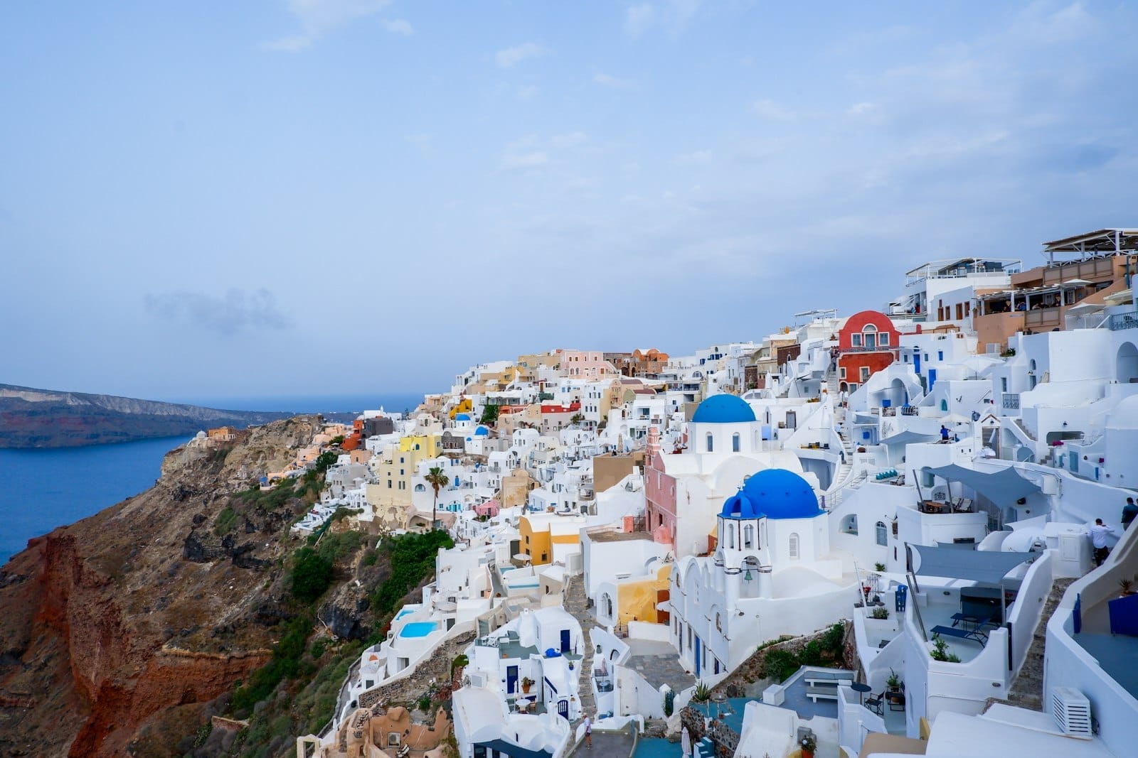 Vista ampla das icônicas construções brancas de Santorini com cúpulas azuis sobre o Mar Egeu azul-profundo e falésias dramáticas sob um céu parcialmente nublado.
