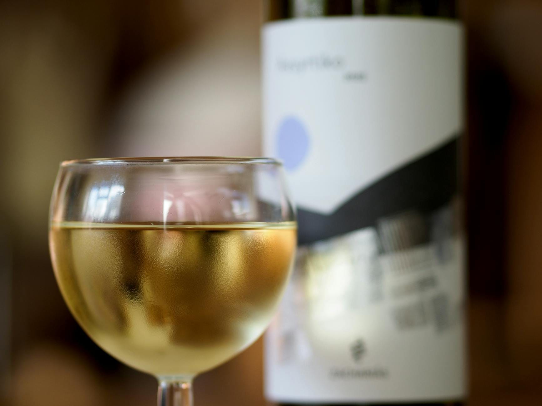 Close-up de uma taça de vinho branco com uma garrafa ao fundo, evocando a atmosfera de uma degustação de vinhos gregos.