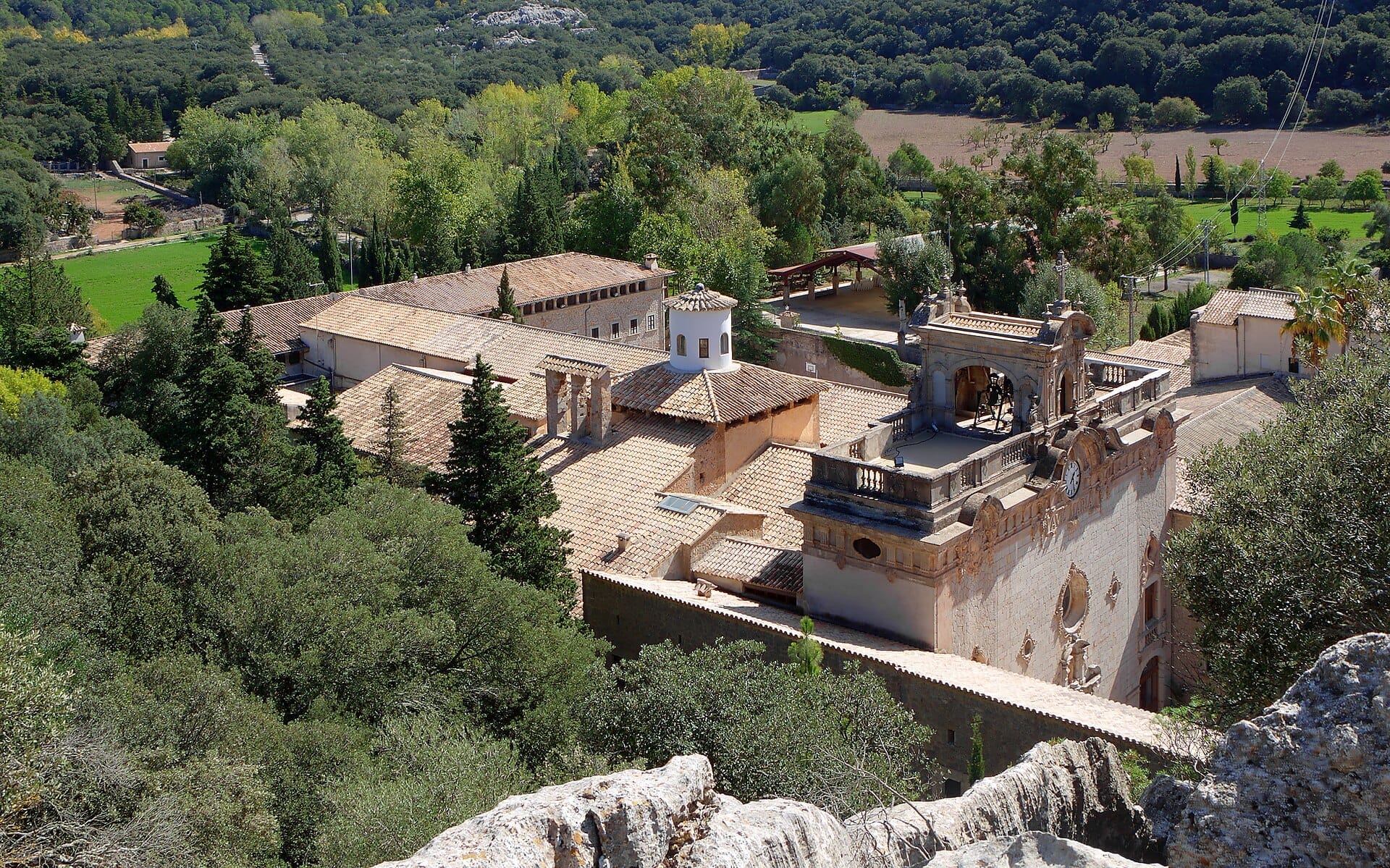 Vista aerea del Santuari de Lluc, con tetti in terracotta, torre dell'orologio e rigogliosa foresta verde nelle montagne della Tramuntana.