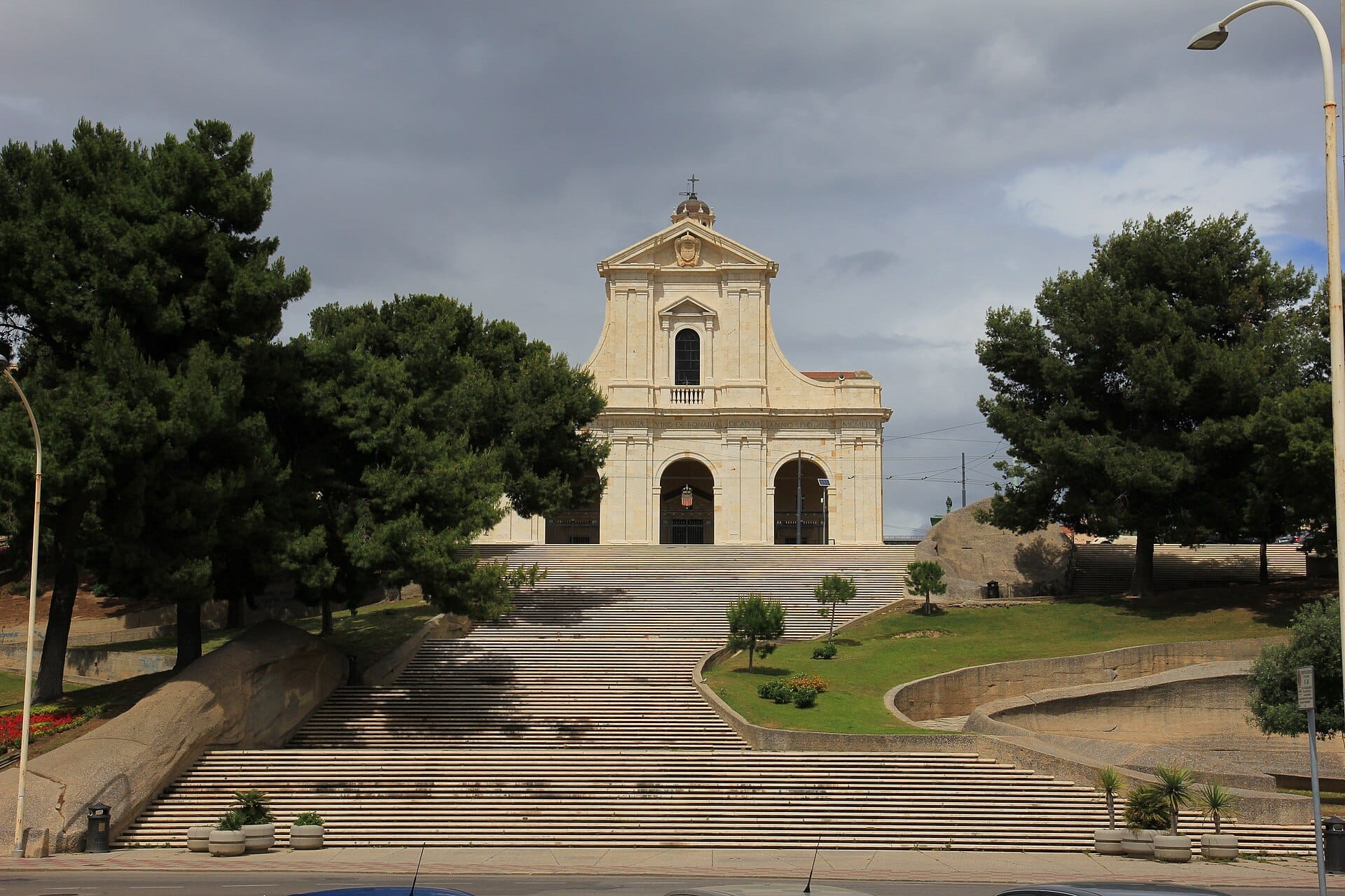 Santuario di Nostra Signora di Bonaria mit seiner großen Treppe, umgeben von grünen Bäumen und dramatisch bewölktem Himmel in Cagliari.