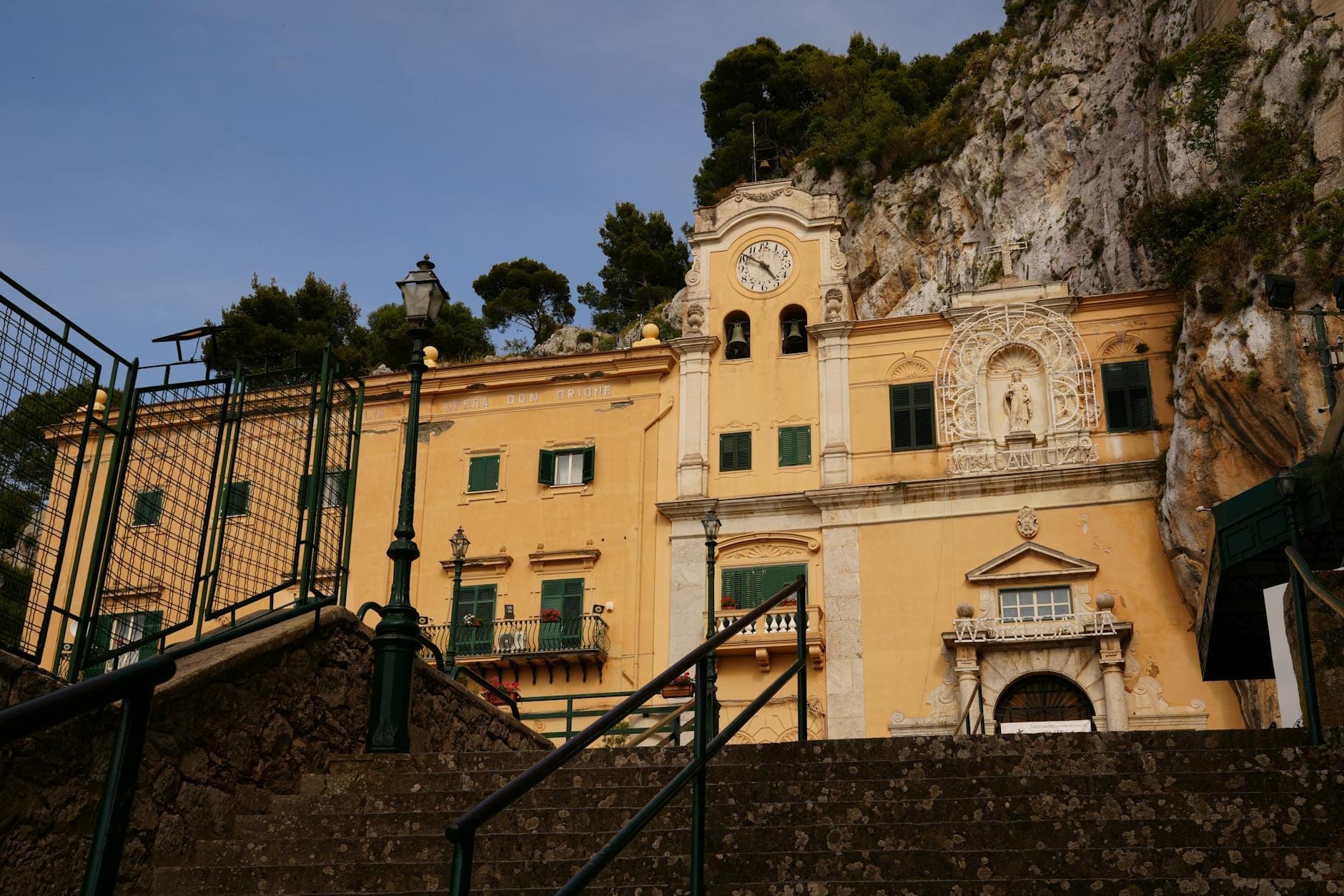 Treppenaufgang zum in die Felswand des Monte Pellegrino eingelassenen Heiligtum der Heiligen Rosalia bei Palermo, mit gelber Fassade, Glockenturm und landschaftlich reizvoller Umgebung.