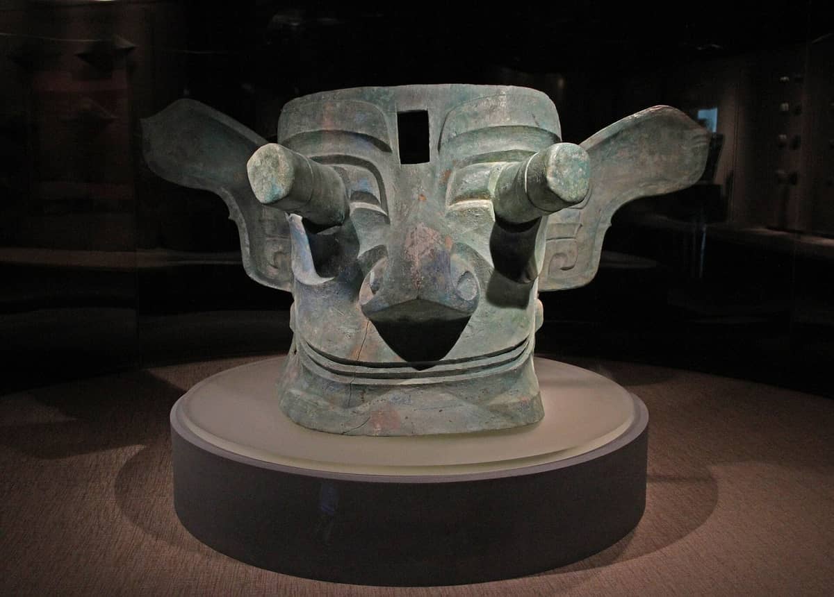 Eine riesige antike Bronzemaske mit großen hervorstehenden Augen, ausgestellt auf einem runden Sockel im Sanxingdui-Museum unter dramatischer Beleuchtung.