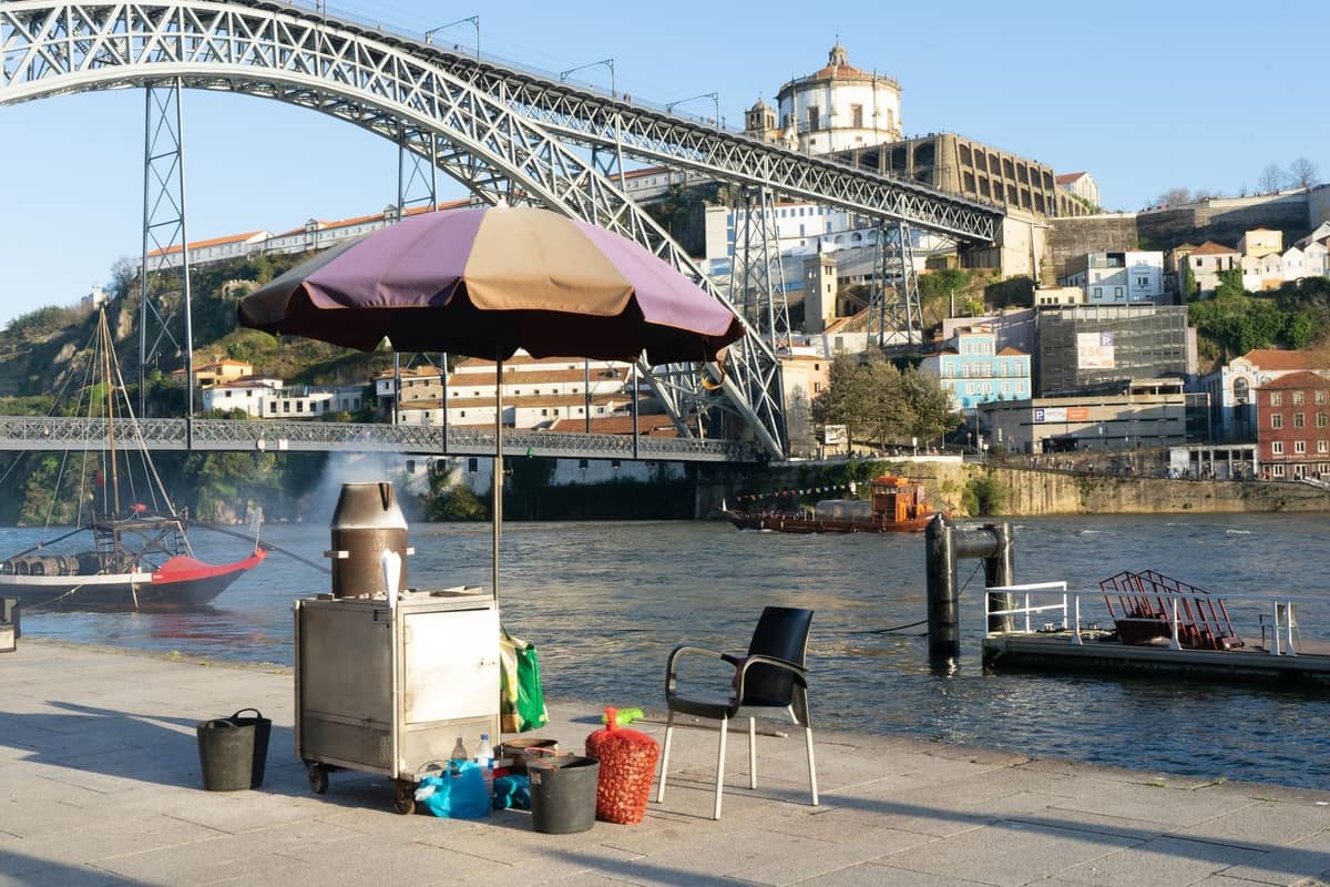 Szene am Flussufer in Porto mit einem Straßenimbisswagen und Grill am Douro, mit der ikonischen Brücke und der historischen Stadt im Hintergrund.