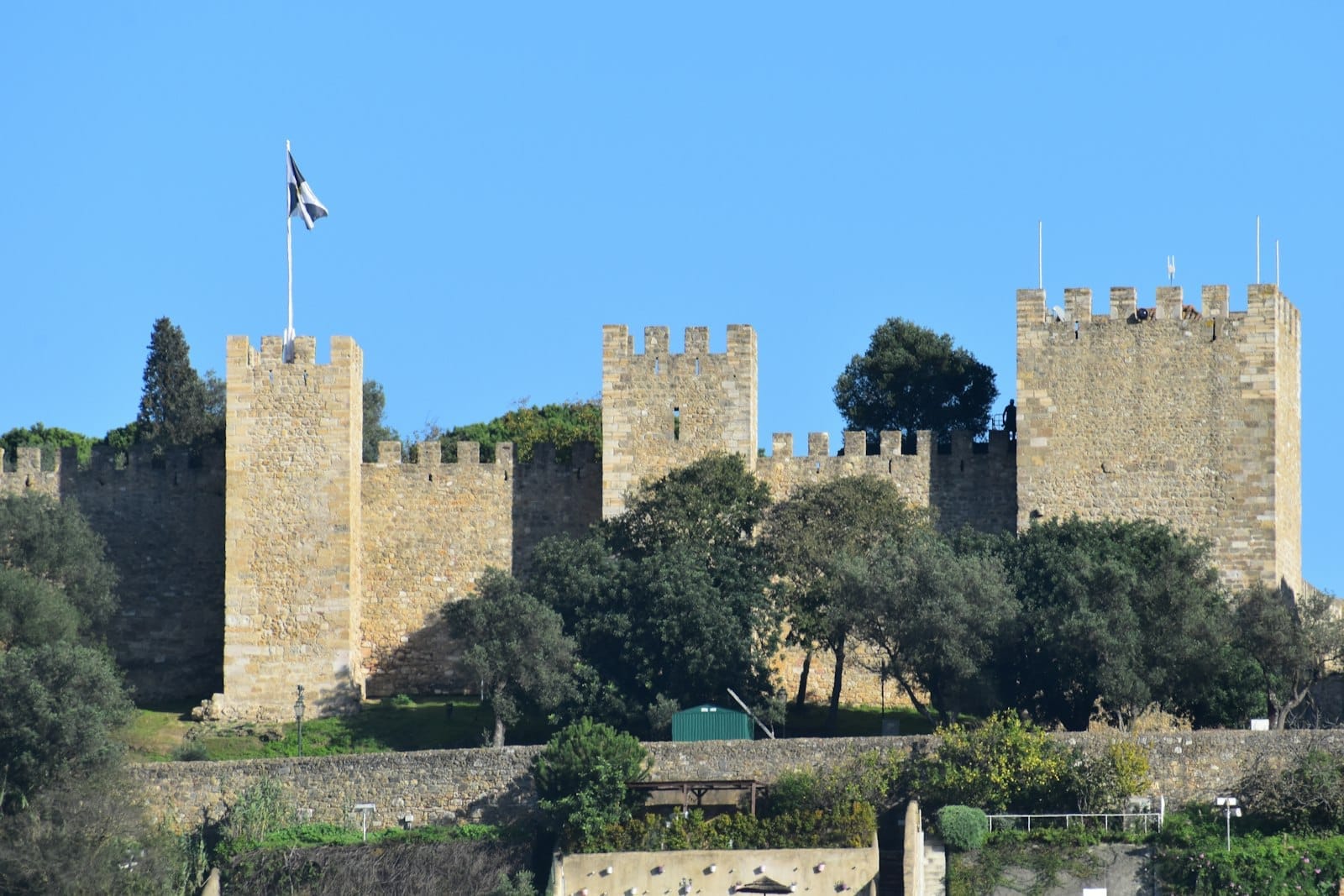 Torri in pietra e mura fortificate del Castello di São Jorge si innalzano sopra alberi verdi sotto un cielo azzurro limpido a Lisbona, Portogallo.