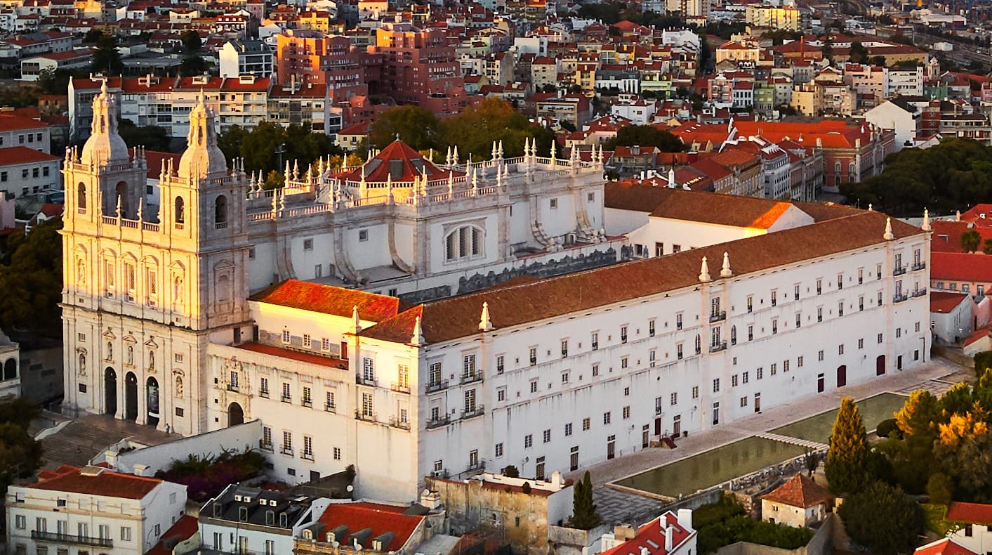 Vista aerea della Igreja de São Vicente de Fora con la sua facciata barocca bianca, il tetto di tegole rosse e il panorama di Lisbona al tramonto.