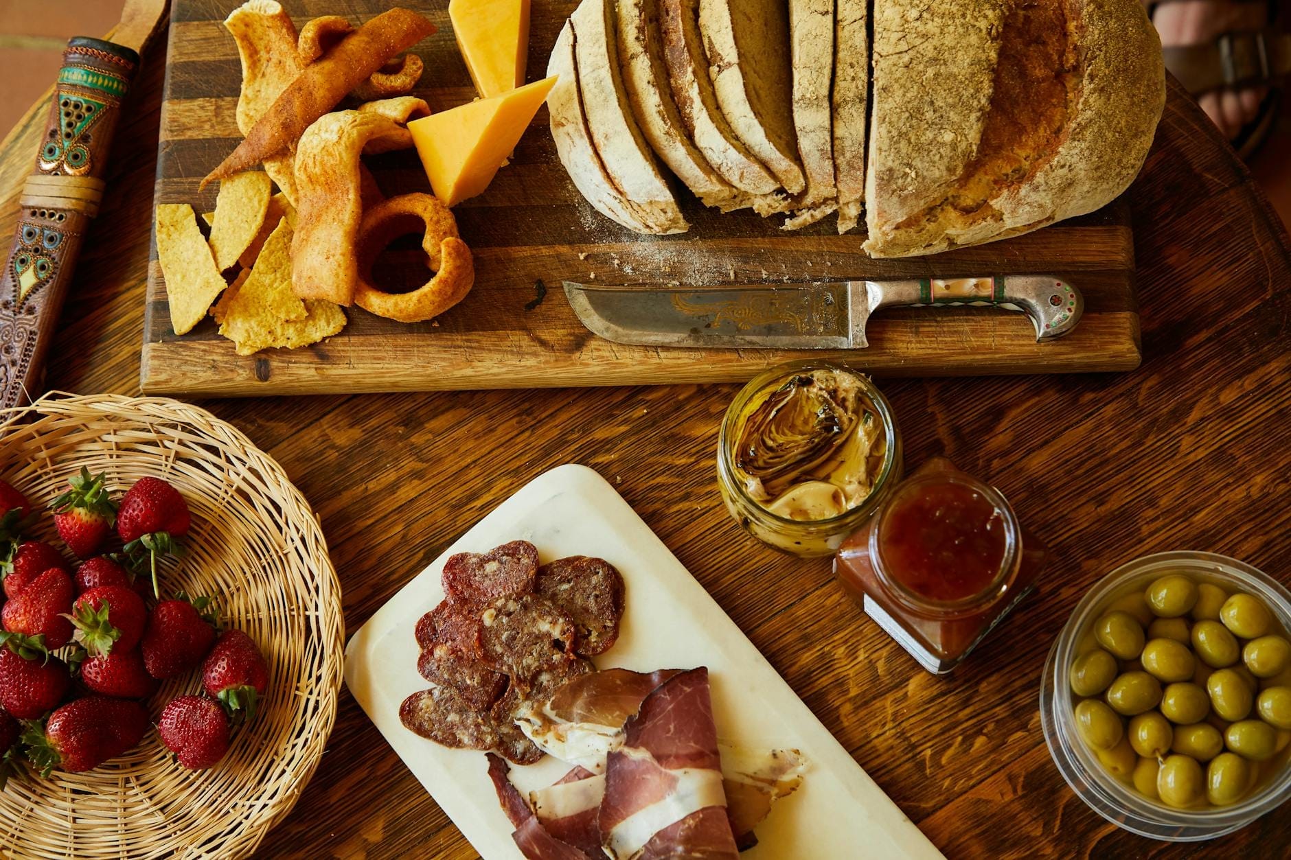 Ein rustikaler Tisch mit geschnittenem Brot, Käse, Aufschnitt, Oliven, Erdbeeren und Eingemachtem, der eine typische sardische Mahlzeit in der Blauen Zone zeigt.