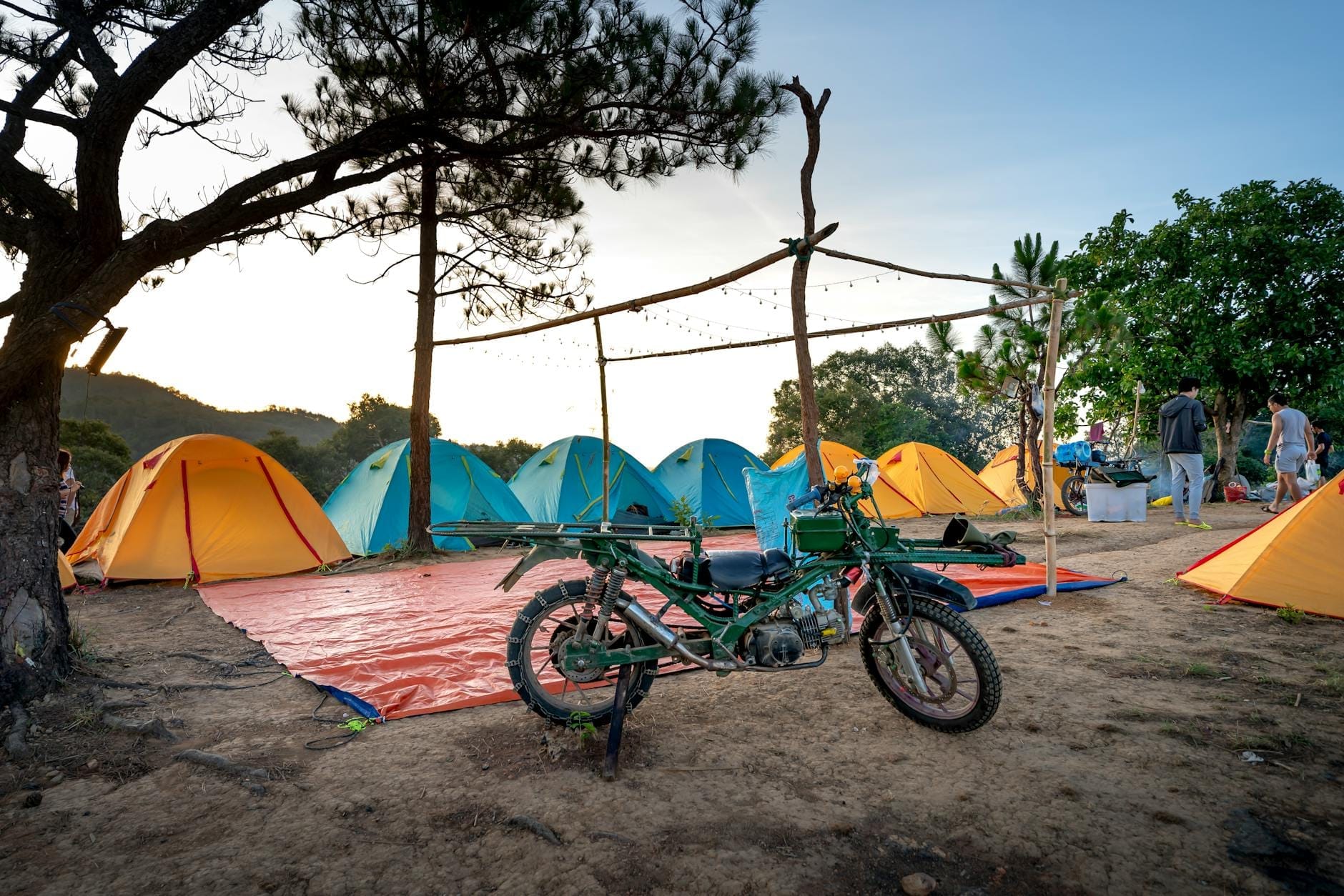 Bunte Zelte unter Kiefern auf einem malerischen Campingplatz, mit einem Motorrad und Menschen im Hintergrund – typisch für das Camping-Abenteuer auf Sardinien.