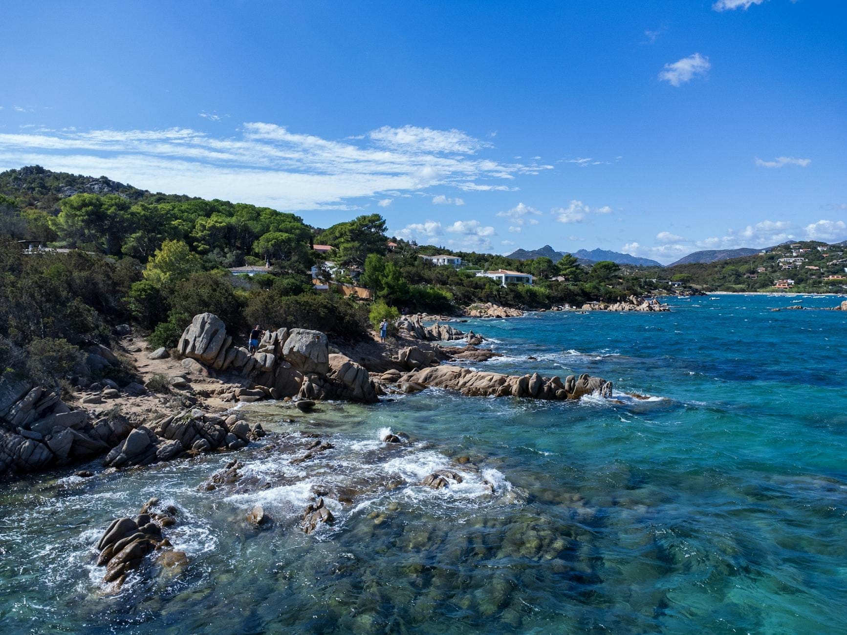 Felsige sardische Küste mit klarem türkisfarbenem Wasser, Pinien und entfernten Siedlungsgebieten im Sonnenschein, ideal zum Campen in der Nähe der Top-Sehenswürdigkeiten.