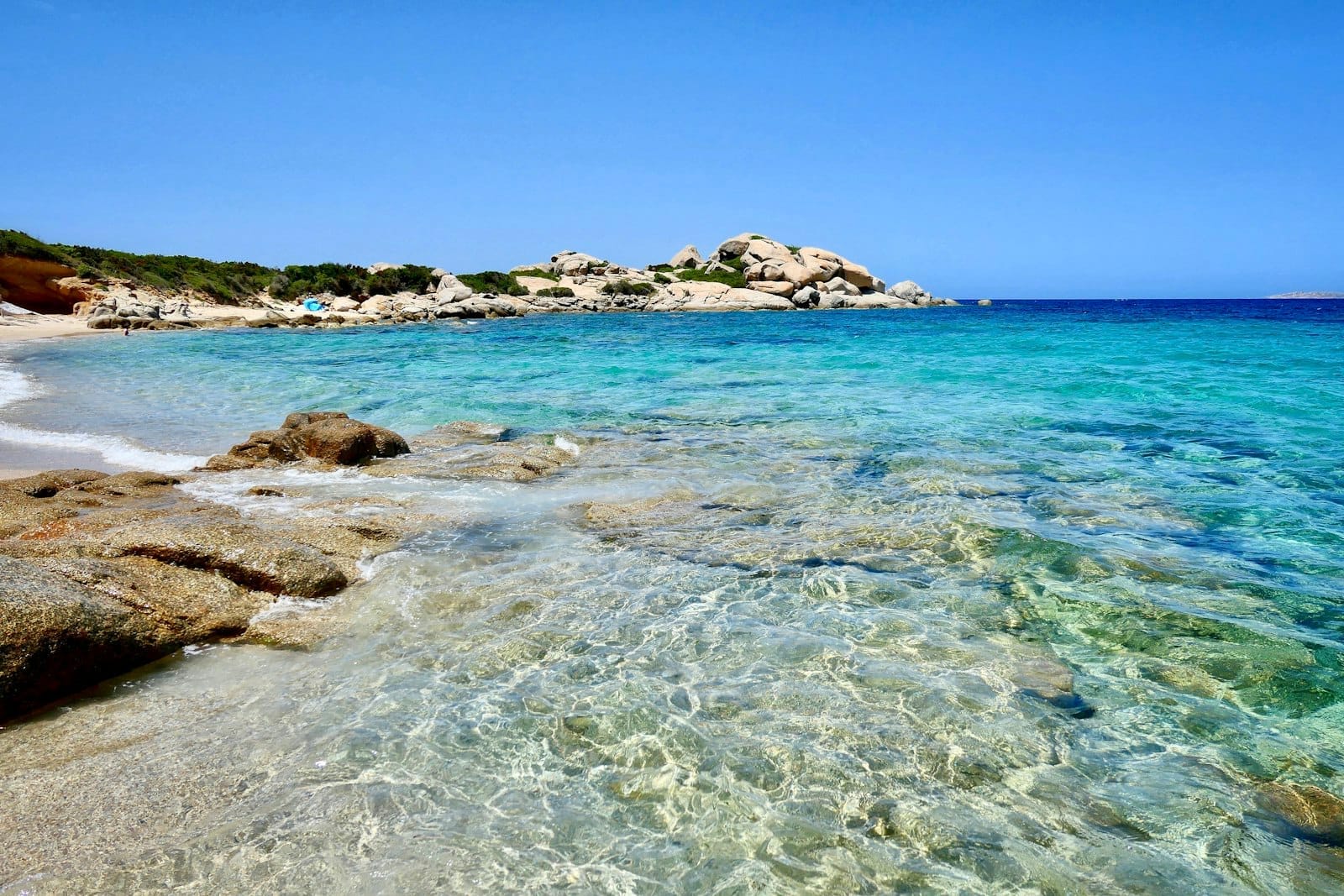 Klares türkisfarbenes Wasser und weißer Sandstrand mit Granitfelsen an der Küste Sardiniens unter einem strahlend blauen Himmel.