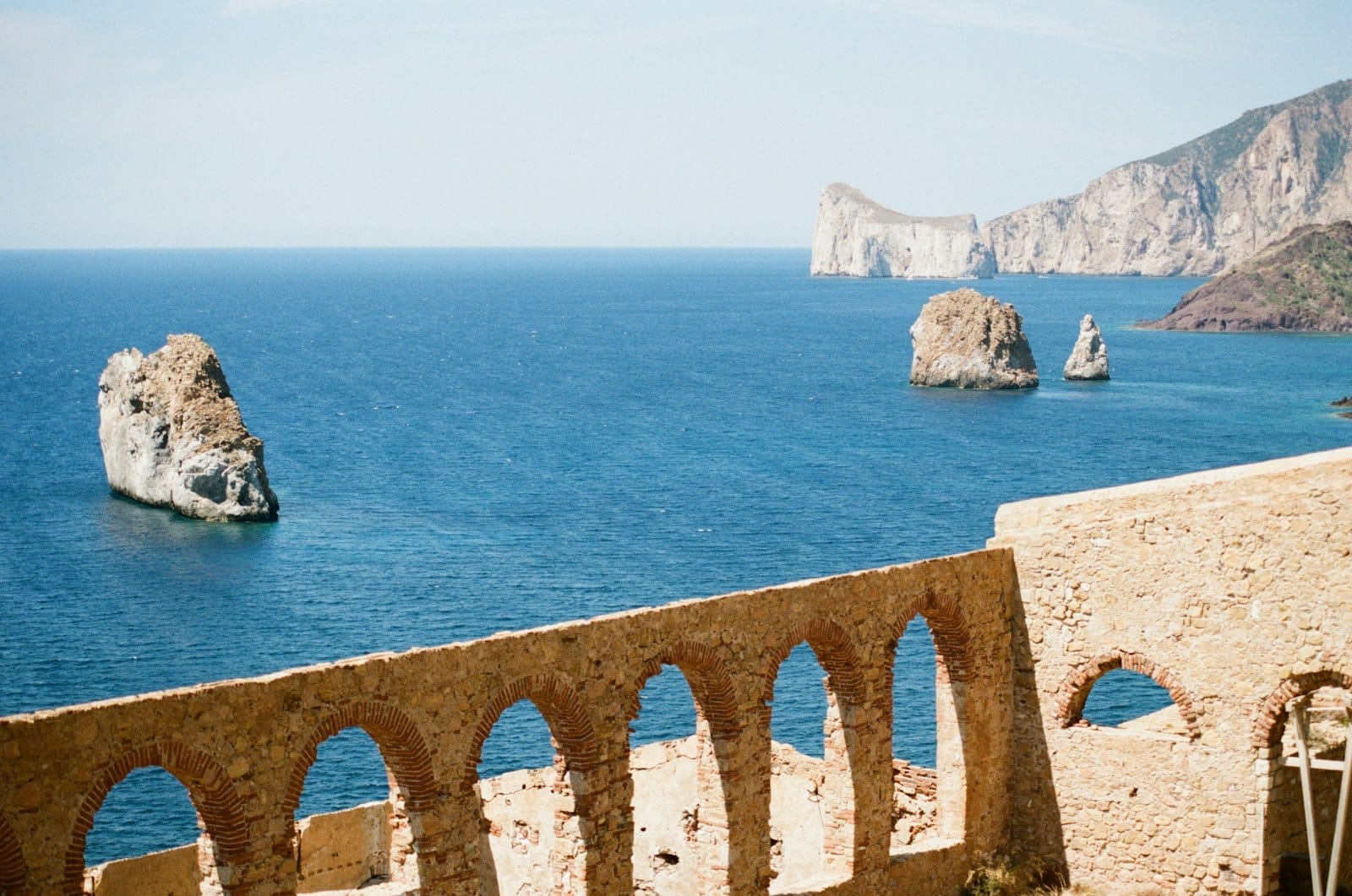 Ruinen eines historischen Steingebäudes mit Bögen mit Blick auf das Meer und dramatischen Felsformationen an der Küste Sardiniens.