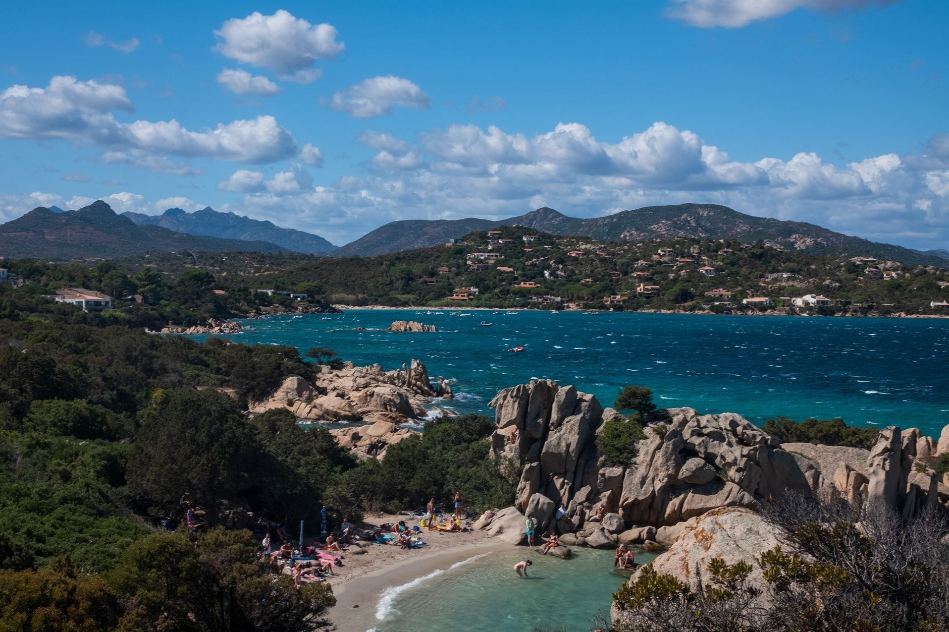 Panoramablick auf die sardische Küste mit Felsformationen, Sandstrand, türkisblauem Wasser und Menschen, die die Sonne genießen – mediterrane Lebensfreude pur.