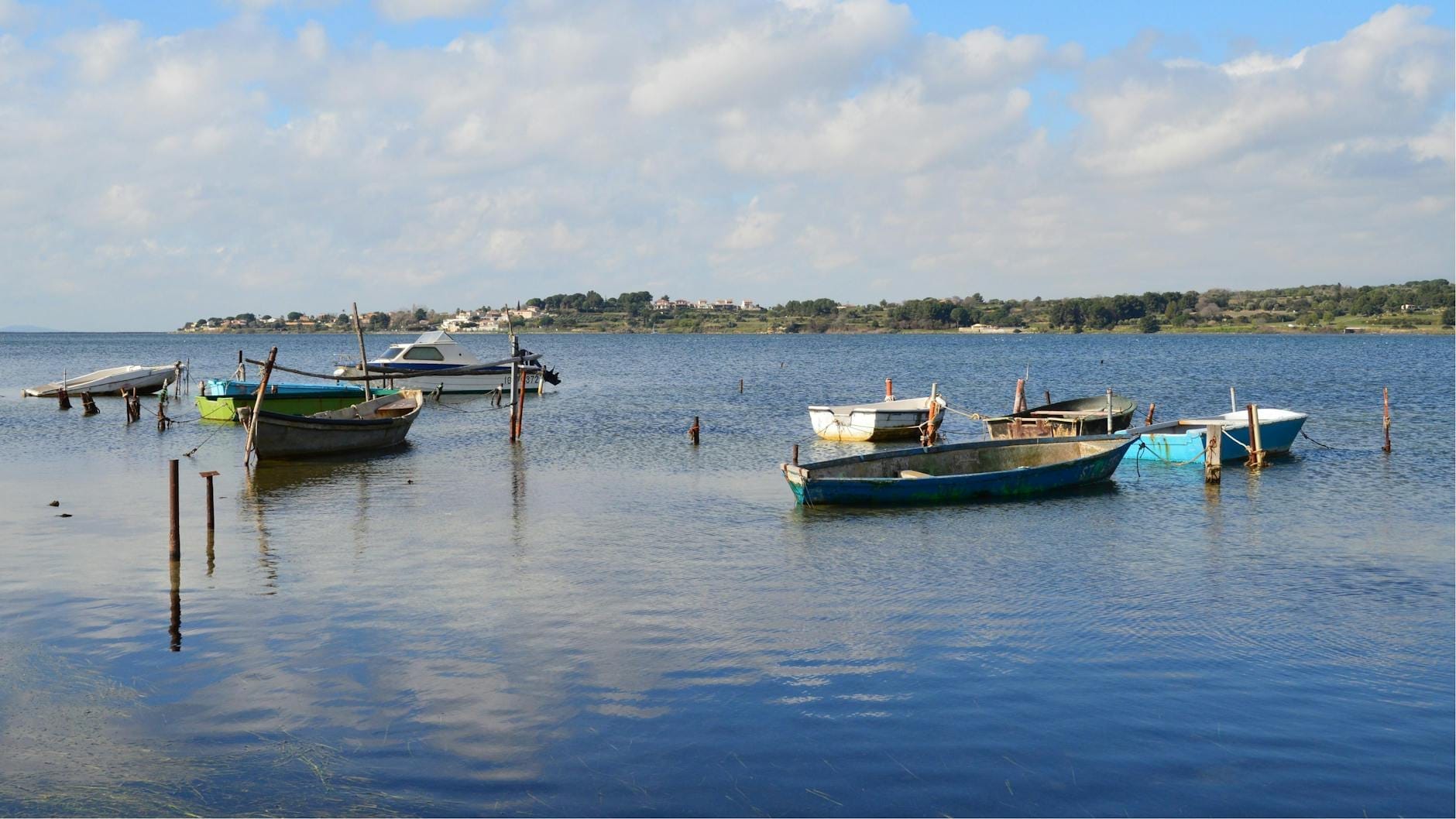 Kleine Fischerboote, die in einer ruhigen Lagune vor Anker liegen, mit fernen Hügeln und einem teilweise bewölkten Himmel, die an die Küstenfischerei Sardiniens erinnern.