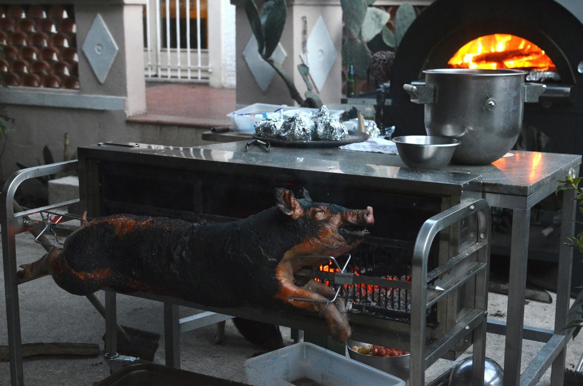 Ganzes Spanferkel, das im Freien am Spieß über offenen Kohlen rotiert, mit Feuerofen im Hintergrund. Traditionelle Fleischzubereitungsszene.