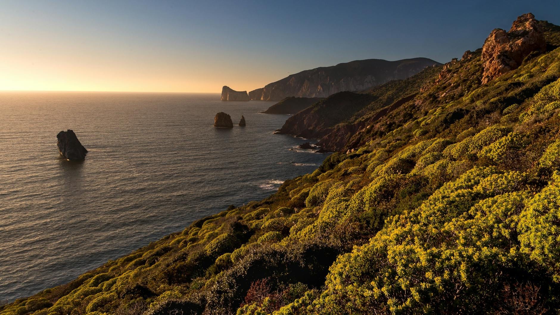 Dramatische Küstenlandschaft Sardiniens bei Sonnenuntergang mit schroffen Klippen, Felsformationen im Meer und üppiger Vegetation über ruhigem blauem Wasser.