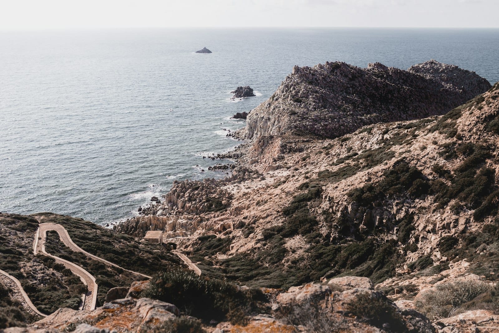 Eine wilde Küste Sardiniens mit gewundenen Pfaden, felsigen Klippen und karger Vegetation mit Blick auf das ruhige Mittelmeer unter einem sanften Himmel.