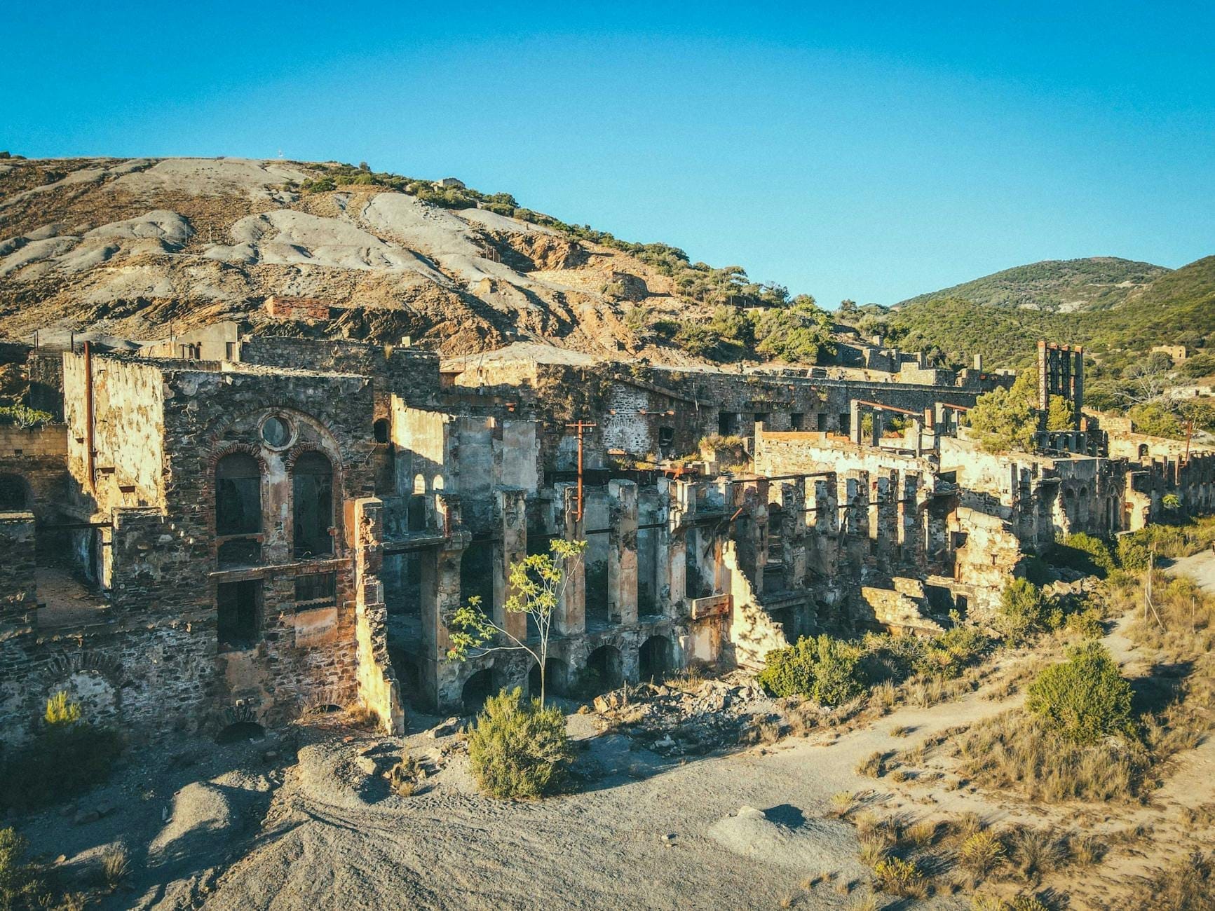 Ruinen eines alten Bergwerkkomplexes in kargen Hügeln, mit beeindruckenden Steingebäuden und natürlicher Landschaft unter blauem Himmel.