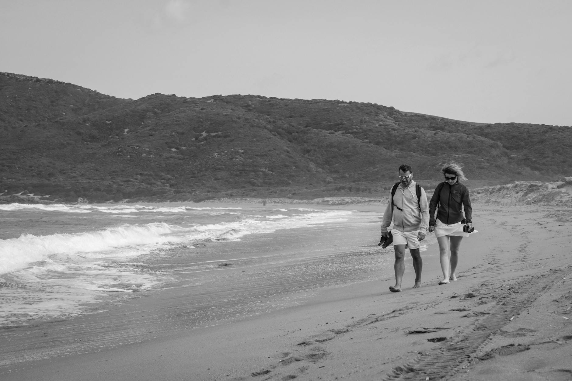 Ein Paar geht Hand in Hand an einem Sandstrand mit sanften Wellen und grünen Hügeln im Hintergrund entlang.
