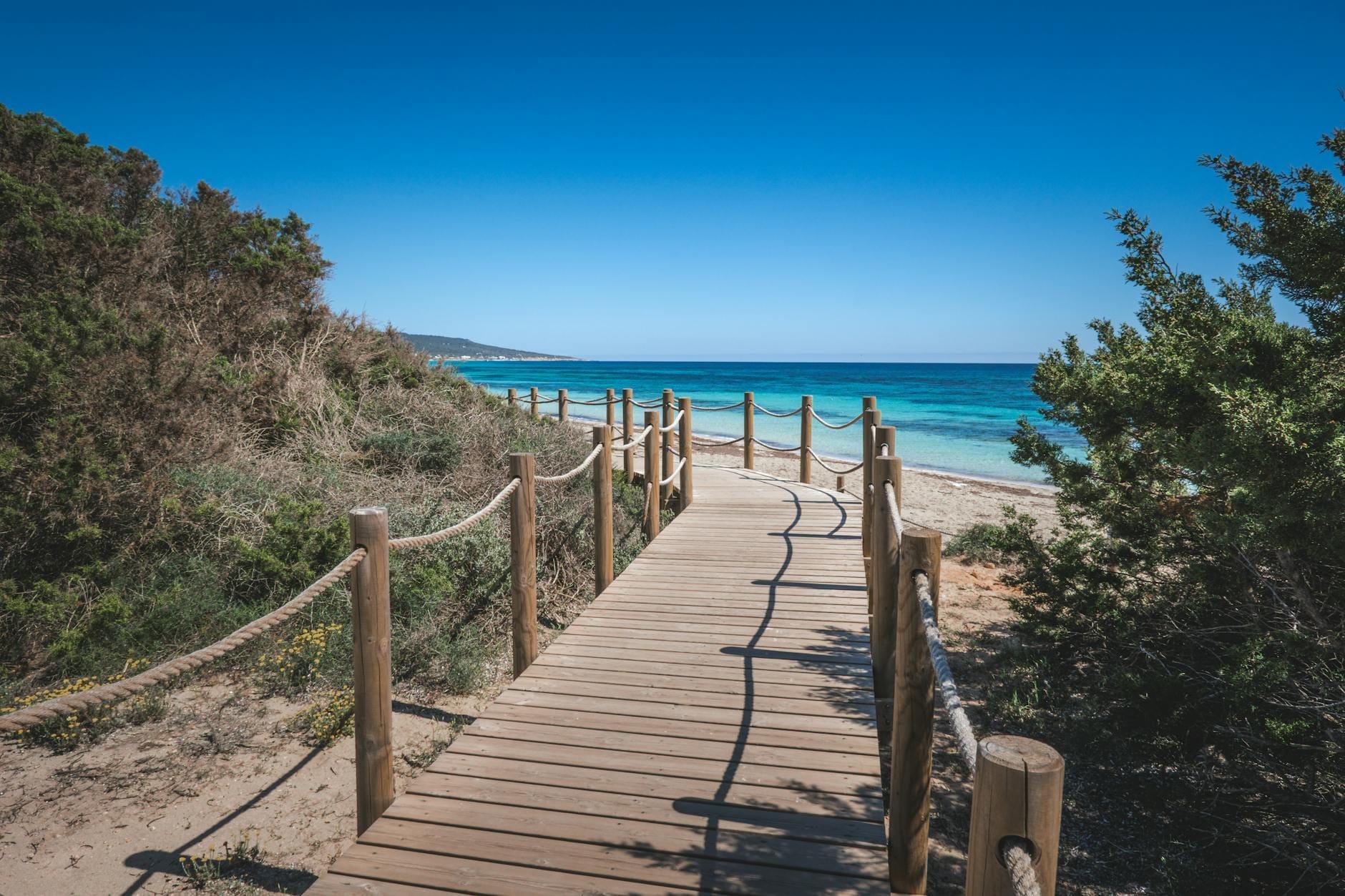 Hölzerner Steg führt zu einem Sandstrand und türkisfarbenem Meer unter einem klaren blauen Himmel, umgeben von grünen mediterranen Sträuchern.