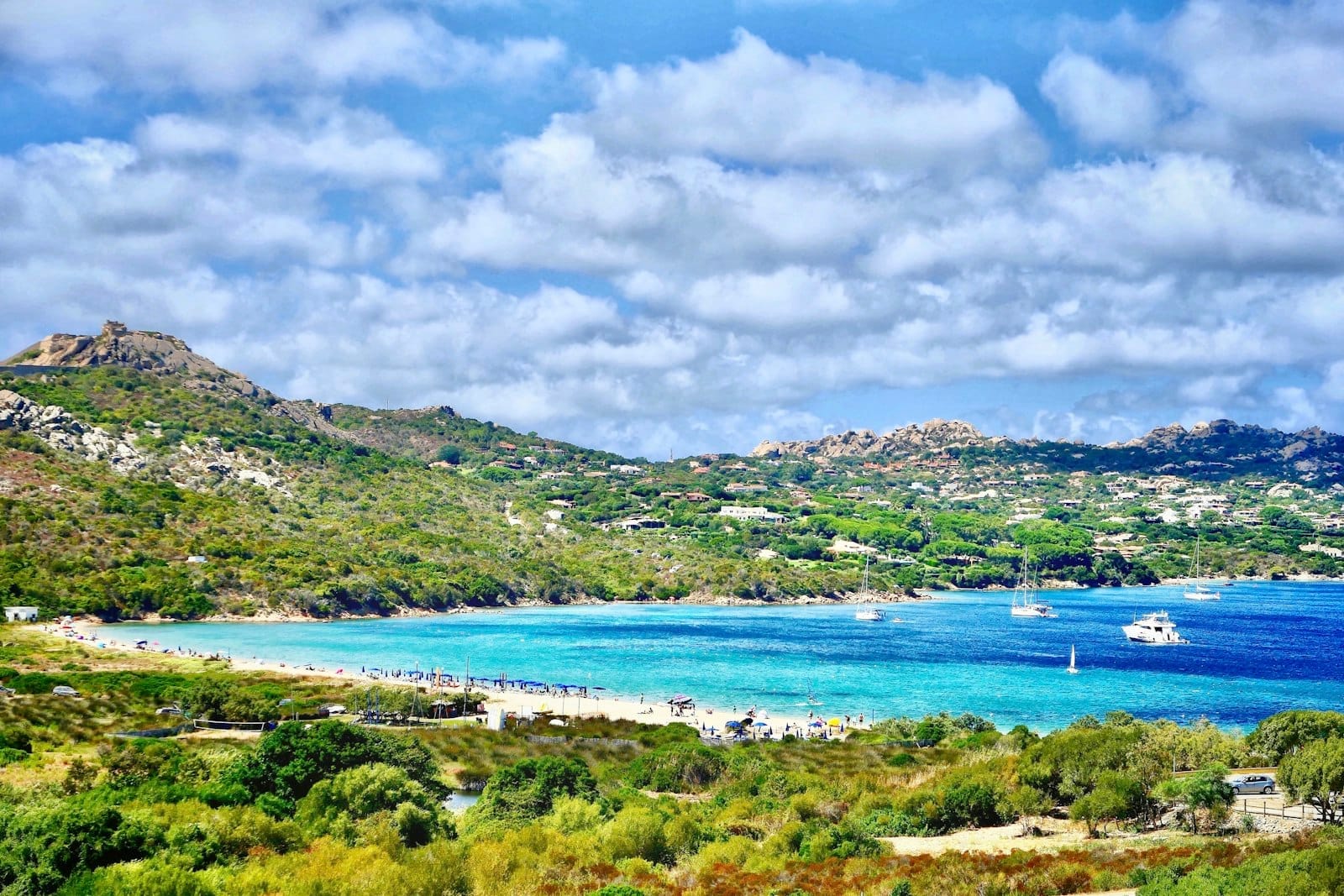 Sonnige sardische Bucht mit türkisfarbenem Wasser, grünen Hügeln, Sandstrand und vereinzelten Booten unter einem leicht bewölkten Himmel im späten Frühling.