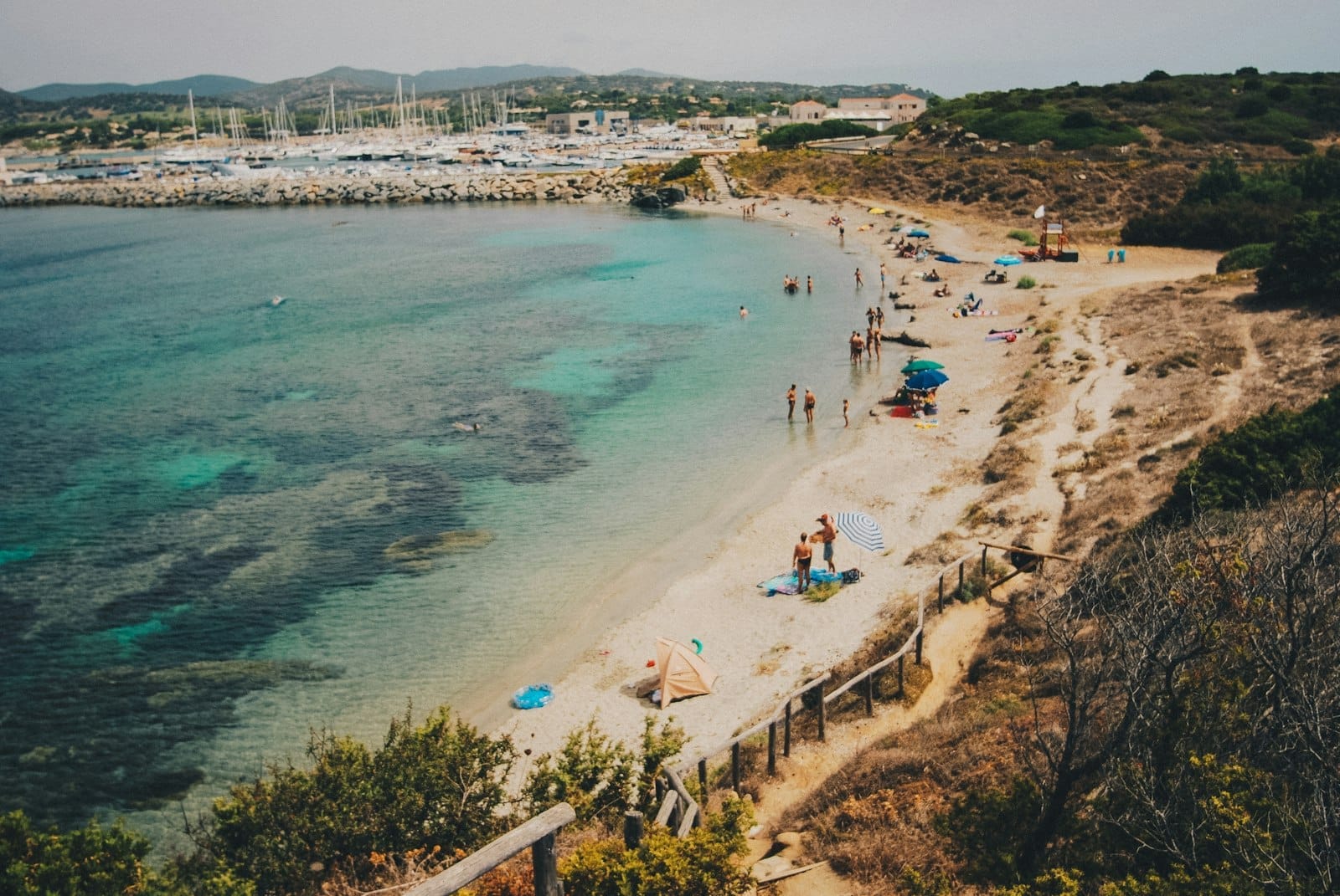 Ein echter sardischer Strand mit vereinzelten Sonnenbadenden und Sonnenschirmen, klarem türkisfarbenem Wasser, sanften Wellen und einem Hafen im Hintergrund unter sonnigem Himmel.