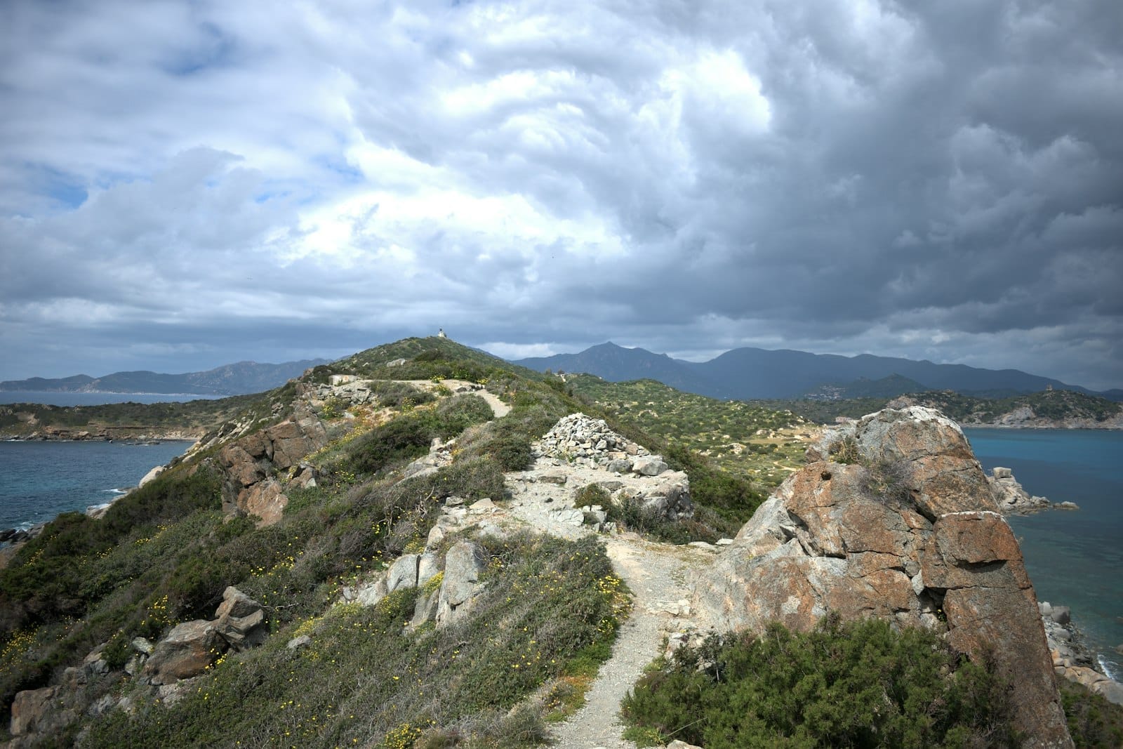 Ein unwegsamer Küstenwanderweg auf felsigen Hügeln in Sardinien, mit dramatischen Wolken, grünen Sträuchern und dem fernen blauen Meer.