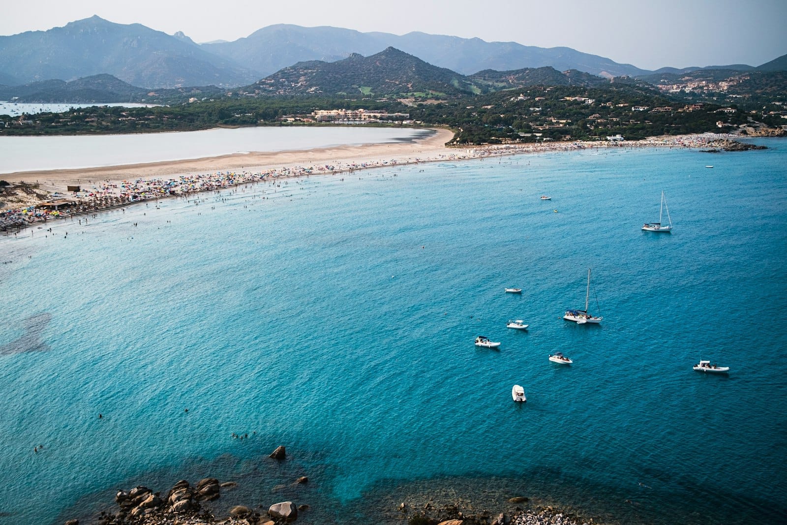 Luftaufnahme eines beliebten sardischen Strandes mit türkisfarbenem Wasser, ankernden Segelbooten und einem belebten Ufer vor Bergen im Sommerlicht.